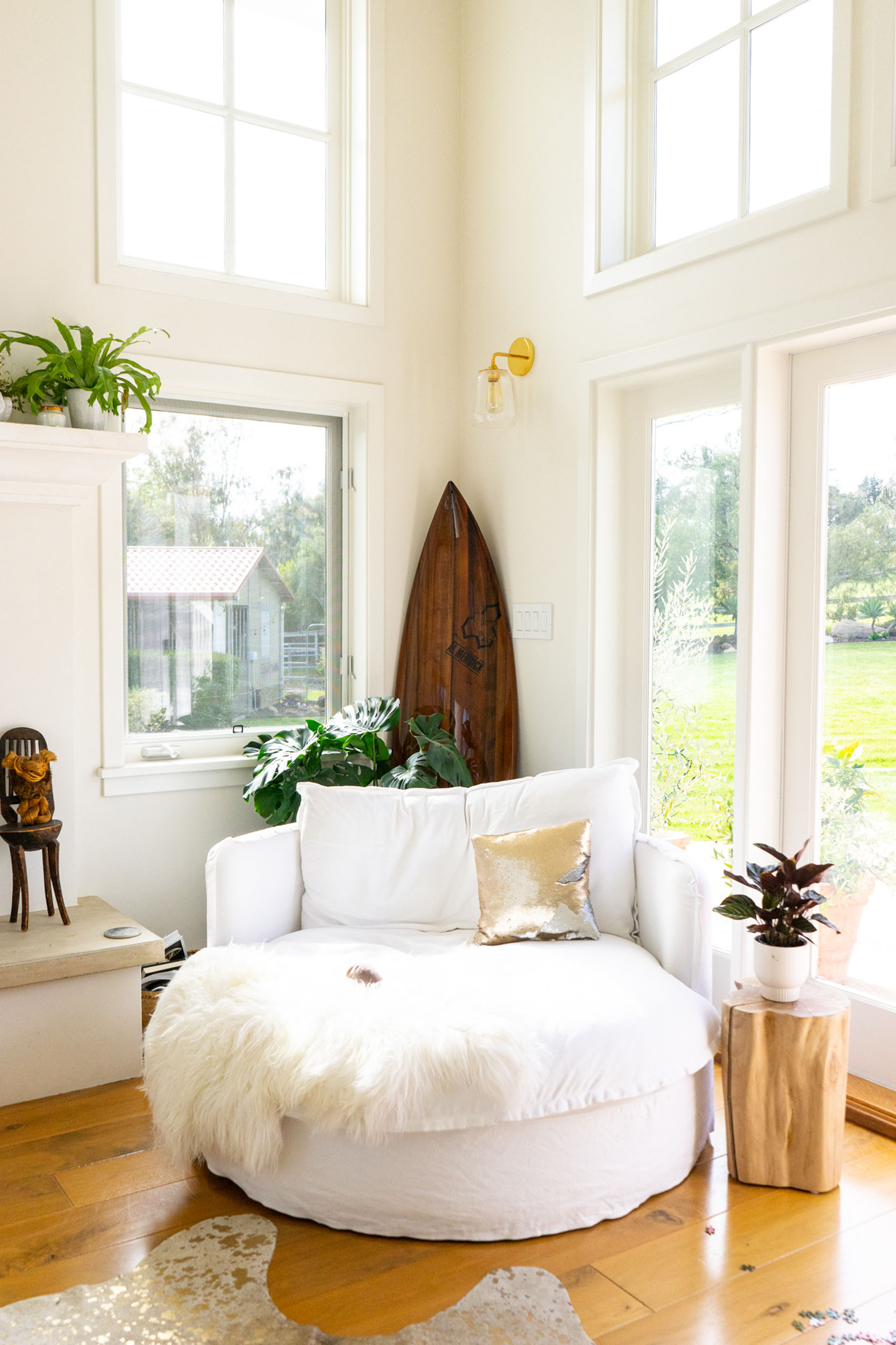 Bright and cozy corner space featuring a large, round white armchair piled with pillows (including a gold sequined one) and a plush white throw blanket. The corner is surrounded by tall windows and transom windows that let in abundant natural light, highlighting a wooden surfboard leaning against the wall, potted green plants, a small wooden side table with a plant on the right, and a glimpse of a fireplace hearth on the left.
