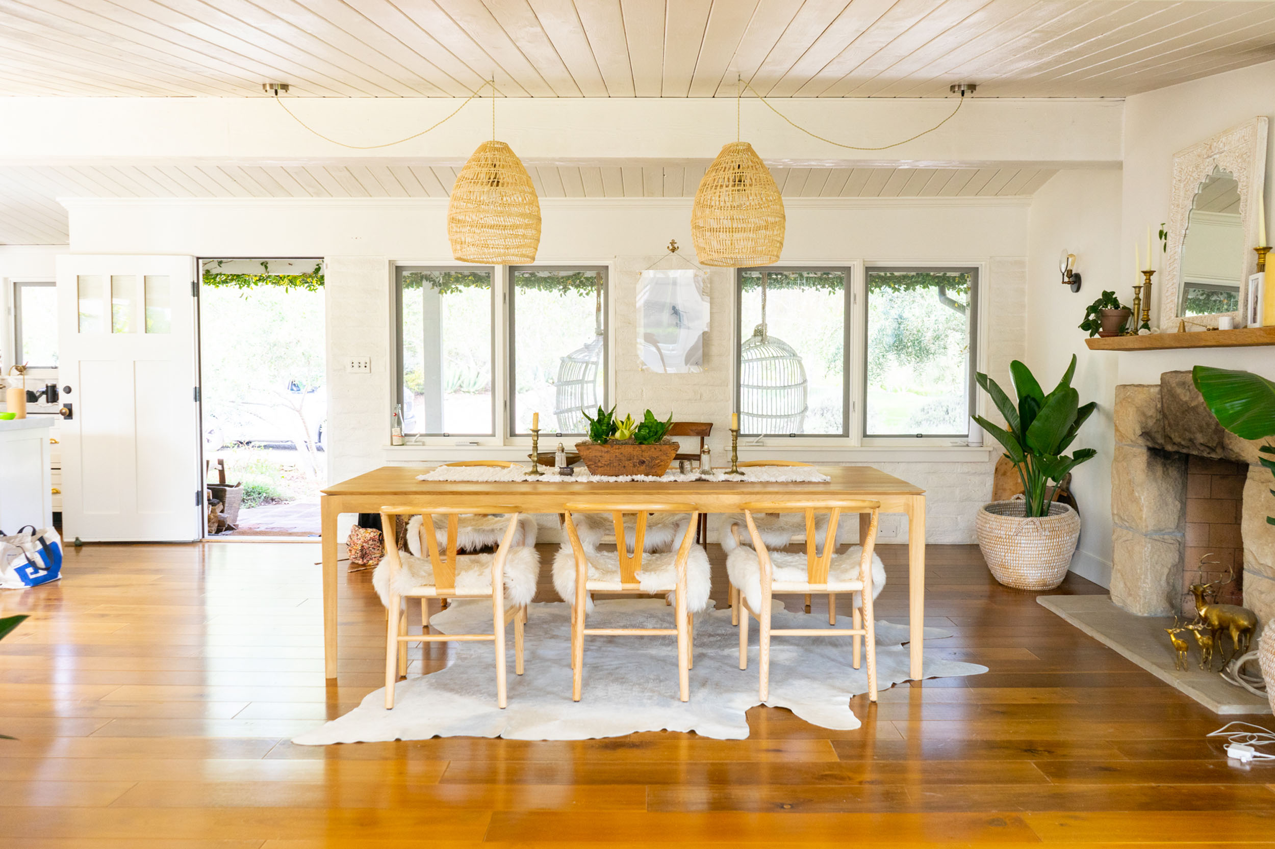Bright and inviting dining and living space featuring a wooden dining table surrounded by wishbone-style chairs with plush sheepskin throws, resting on a cowhide rug. The room has warm wood flooring, white walls, and a row of windows with woven pendant lights hanging above. To the right, a stone fireplace with a wooden mantel features decorative brass accents, complemented by potted greenery and a white door left open to the outdoors on the left.