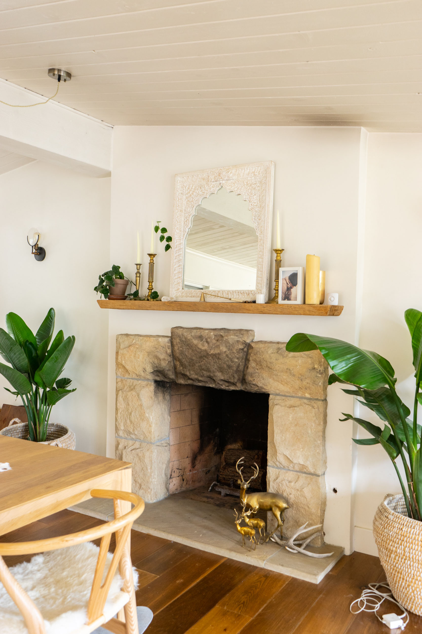 Stone fireplace with a rustic wooden mantel featuring a decorative mirror, brass candlesticks, and candles, flanked by large potted plants, with brass deer figurines and antlers on the hearth.
