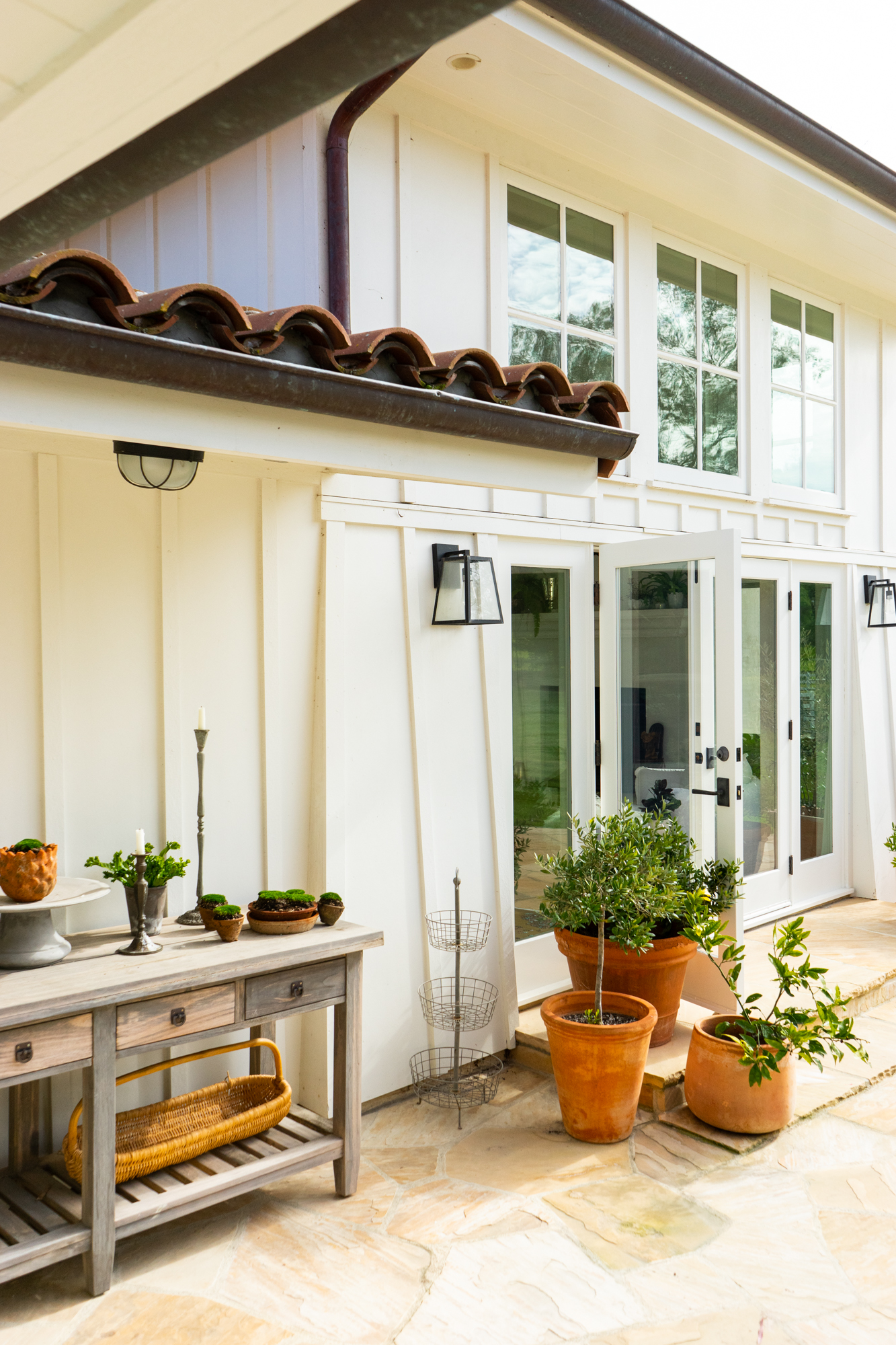 Exterior facade featuring white board-and-batten siding, traditional divided-light windows, and French doors opening onto an irregular flagstone patio. The architecture is accented by a clay barrel tile roof overhang, black metal sconces, a rustic wood console table, and terracotta planters.