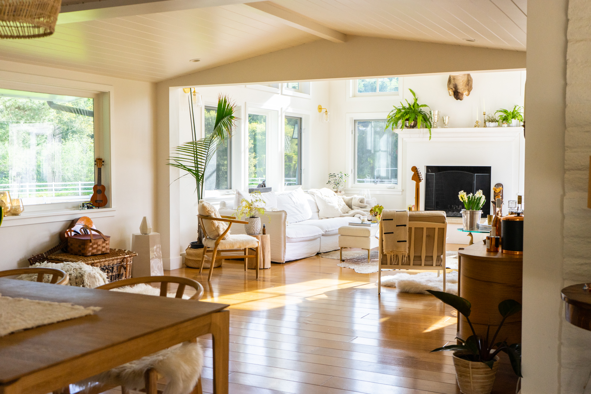 Bright open-plan living and dining space featuring a painted tongue-and-groove vaulted ceiling, expansive picture windows maximizing natural light, and warm hardwood flooring. The transitional interior is anchored by a white fireplace mantel and styled with organic textures, indoor greenery, and eclectic furnishings.