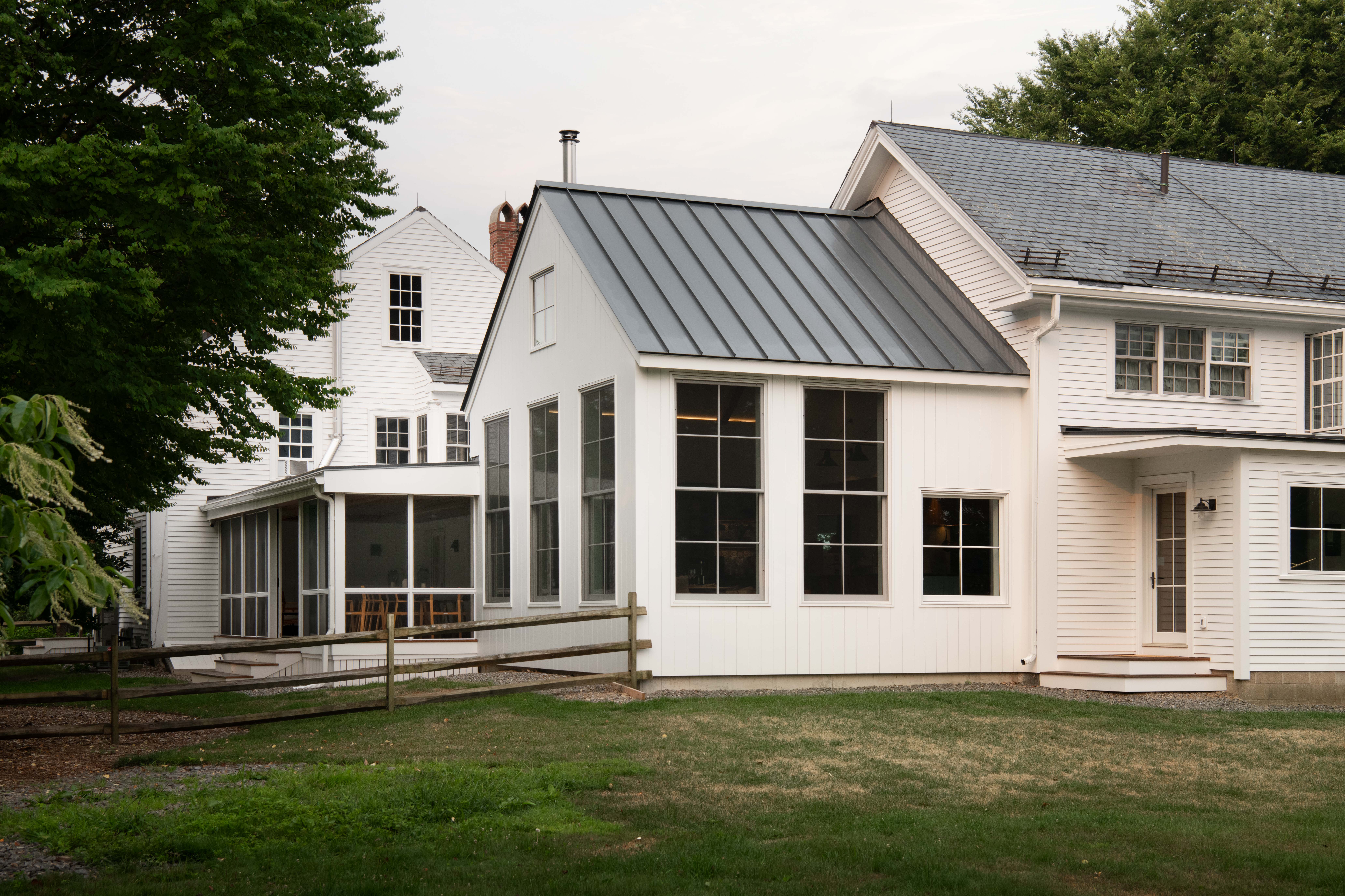 White farmhouse addition with a gray standing seam metal roof, large divided-light windows, and a screened porch, bordered by a wooden split-rail fence and green lawn