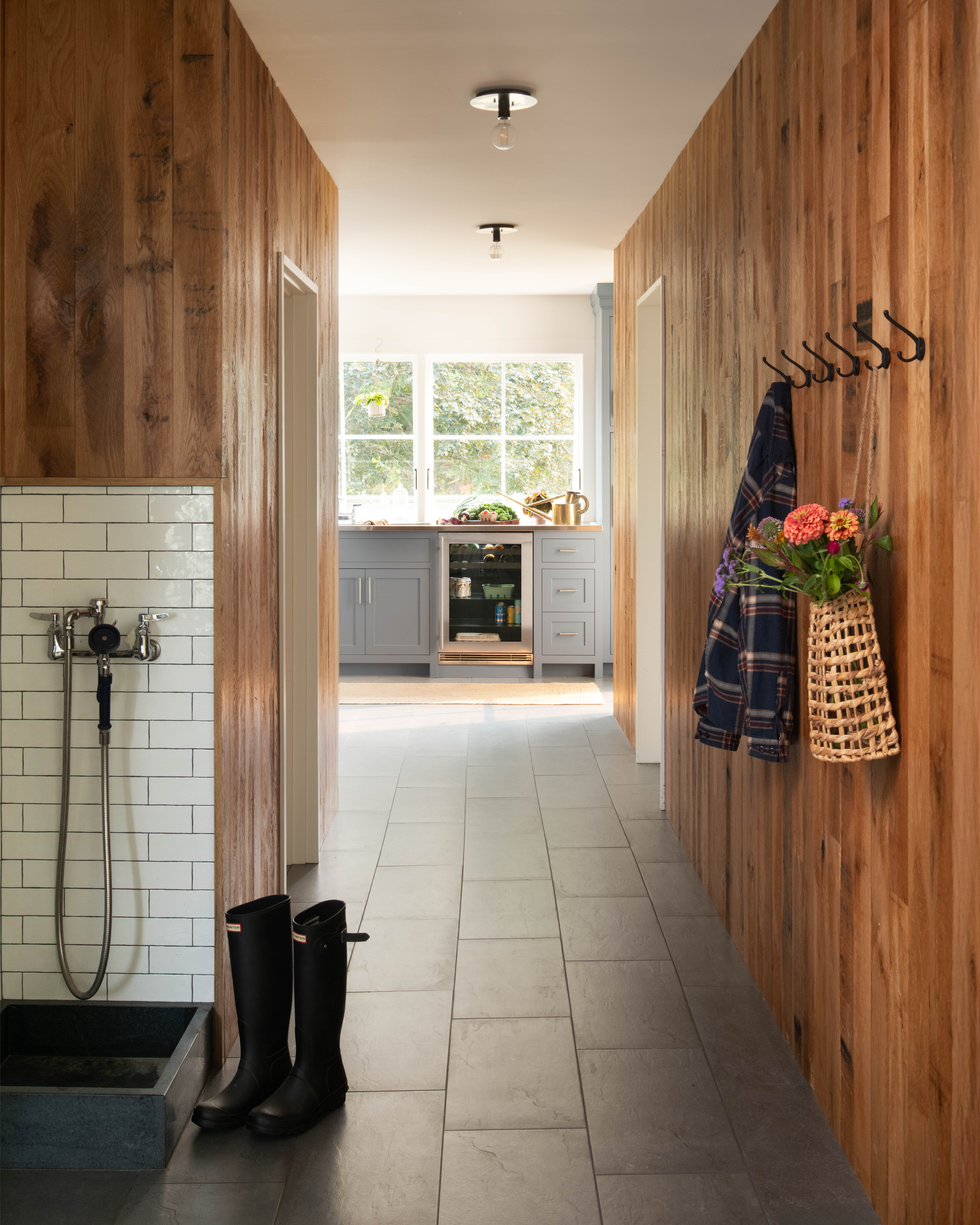 Rustic modern mudroom hallway featuring warm vertical wood-paneled walls, slate tile flooring, a tiled dog washing station with rain shower attachment, and a view into a laundry or prep room with blue cabinetry