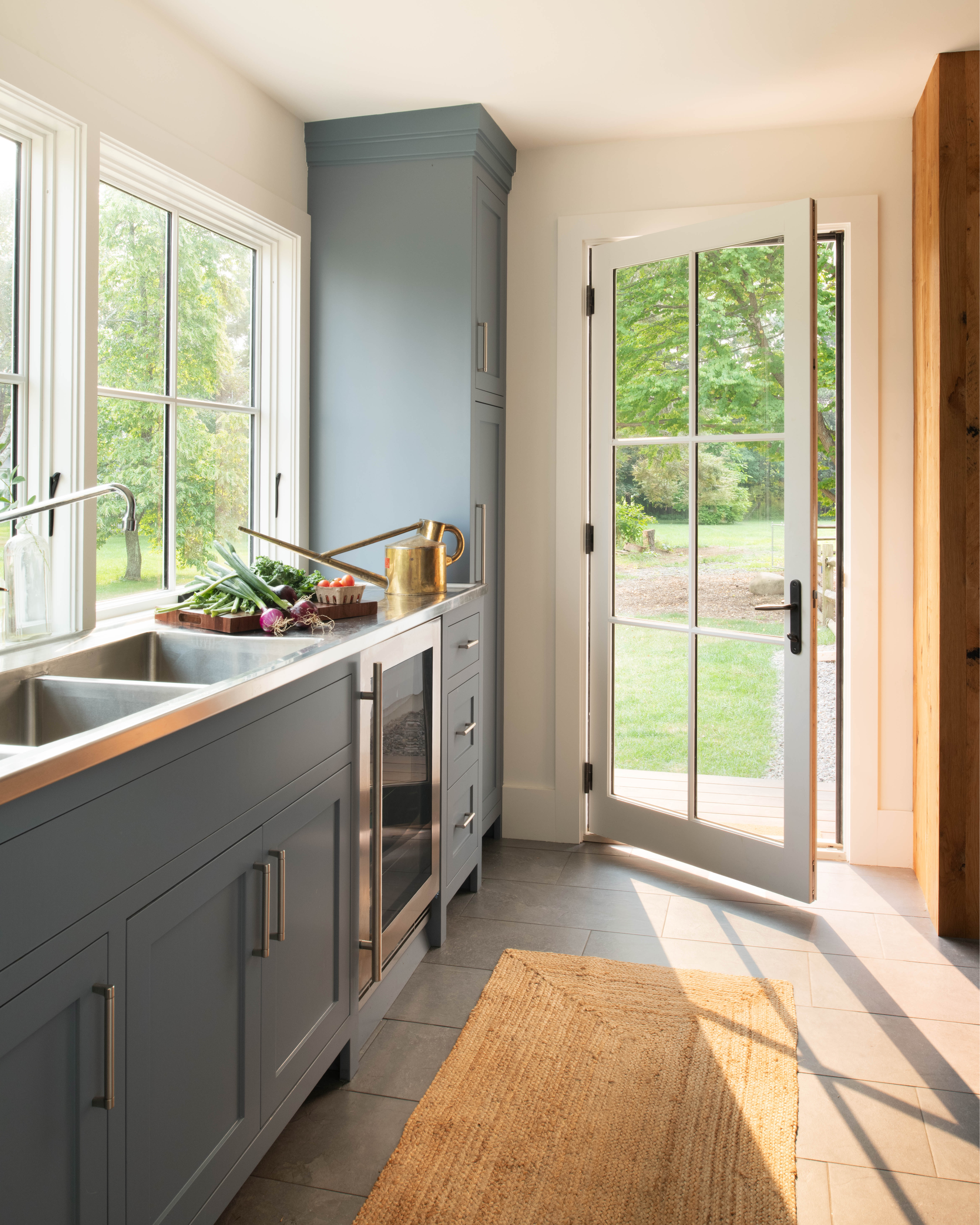 Bright mudroom and prep kitchen featuring slate tile floors, blue shaker cabinetry with a stainless steel sink, beverage cooler, and an open glass-paneled back door leading to the garden