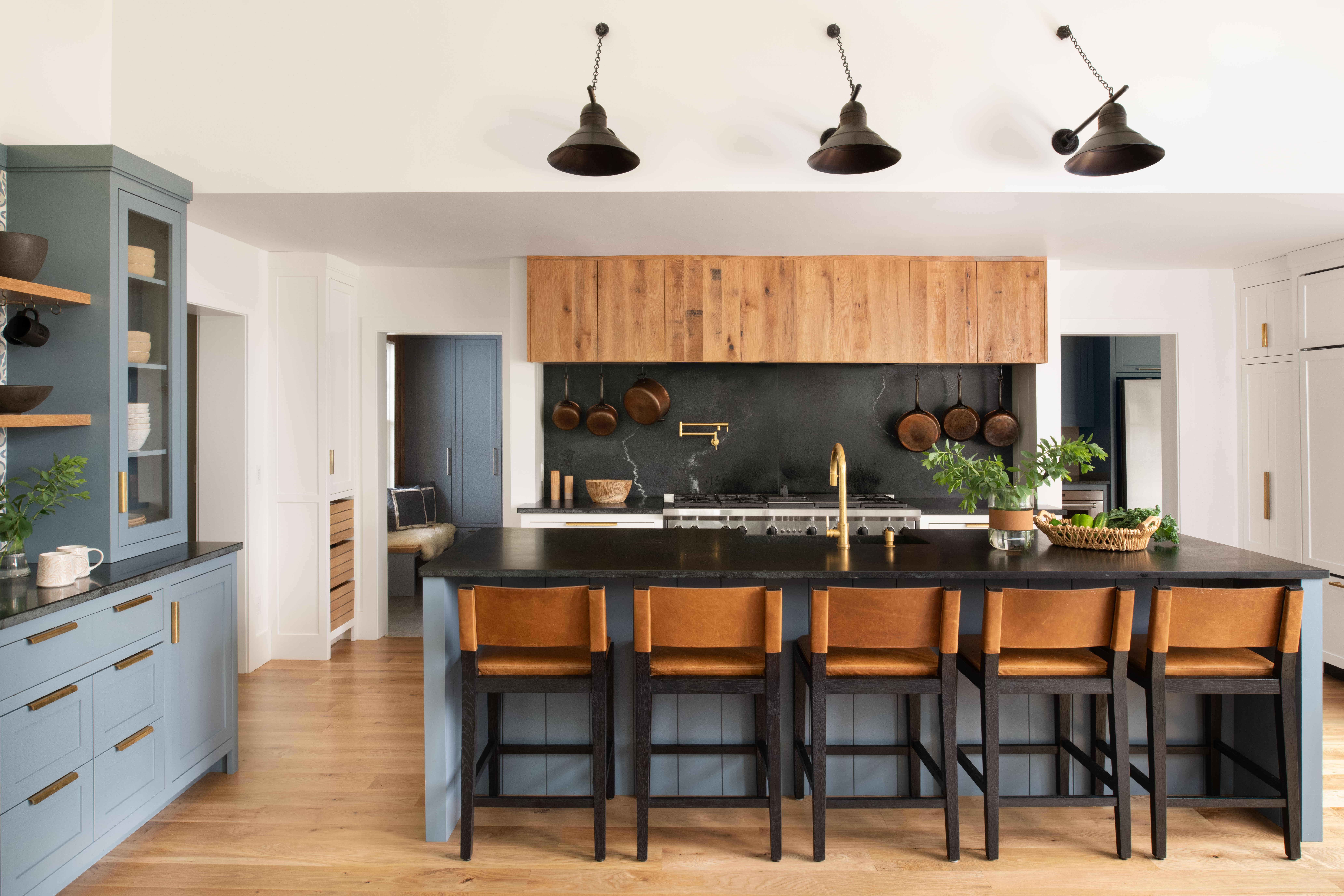 Modern kitchen interior featuring natural wood upper cabinets, a dark soapstone backsplash with brass pot filler, hanging copper cookware, and a blue island with leather barstools