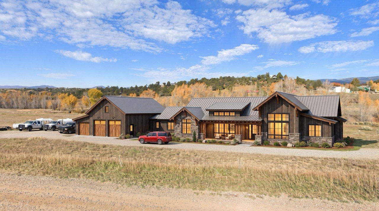 Sprawling modern rustic estate featuring dark board-and-batten siding, a standing seam metal roof, and stone base accents. The expansive home includes large divided-light windows glowing with warm interior light and a multi-bay garage with wood doors, set against a rural landscape with autumn foliage.
