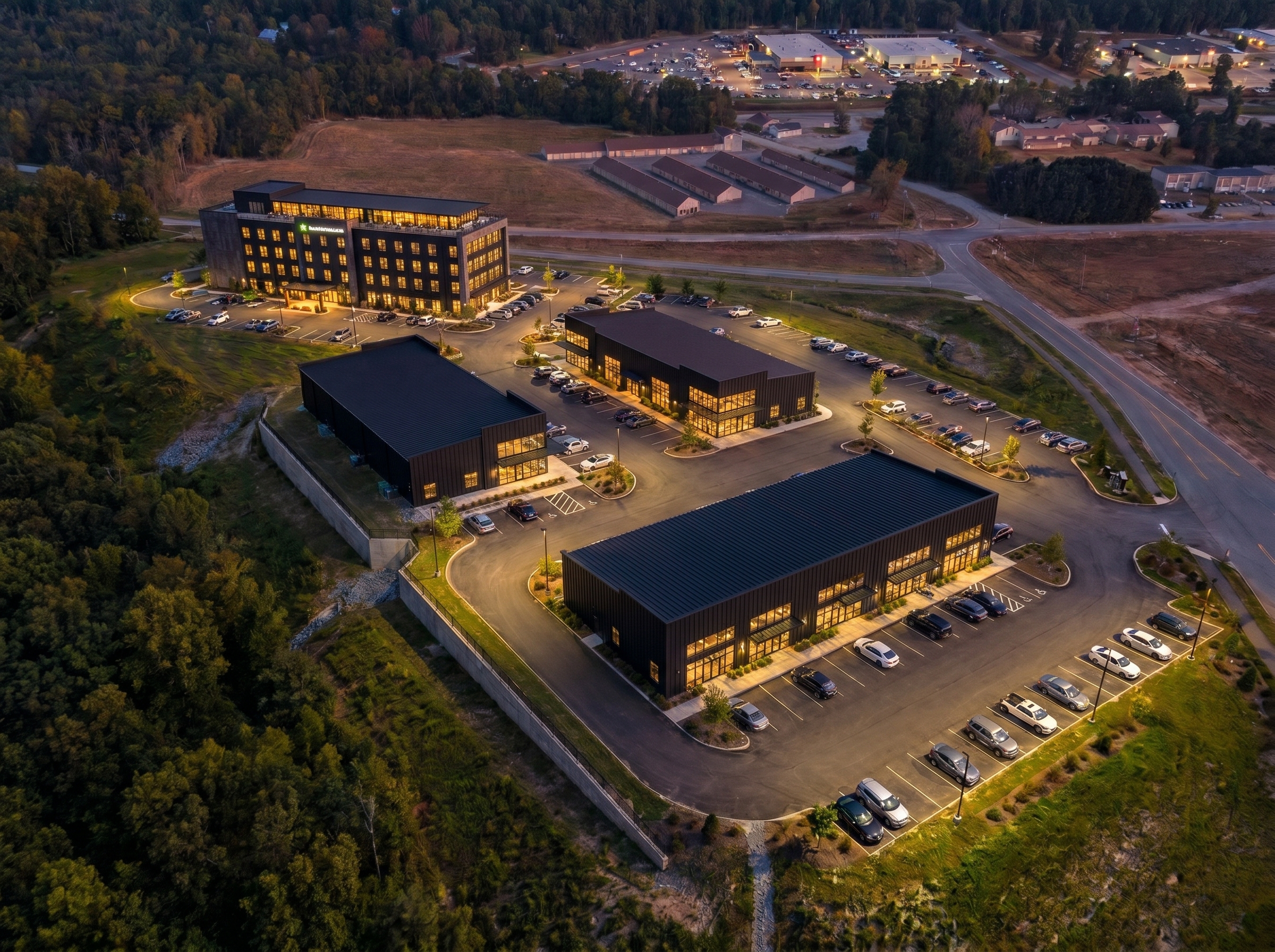 Aerial dusk view of a modern commercial development featuring dark-clad multi-story and single-story retail buildings surrounded by illuminated parking lots and forested landscape