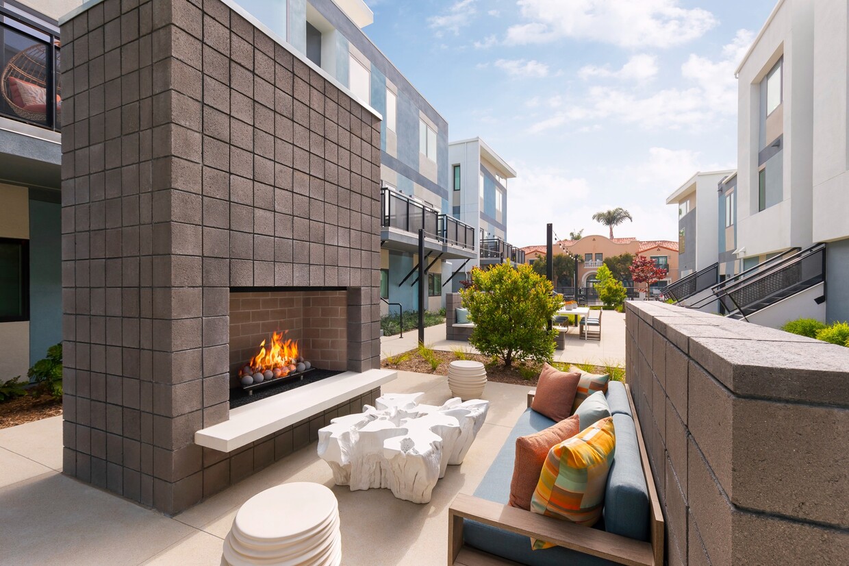 Modern outdoor patio space featuring a block-tile fireplace with a burning fire, a blue sofa with colorful throw pillows, a white carved wood-root coffee table, matching round side tables, and lush landscaping against a multi-story building backdrop.