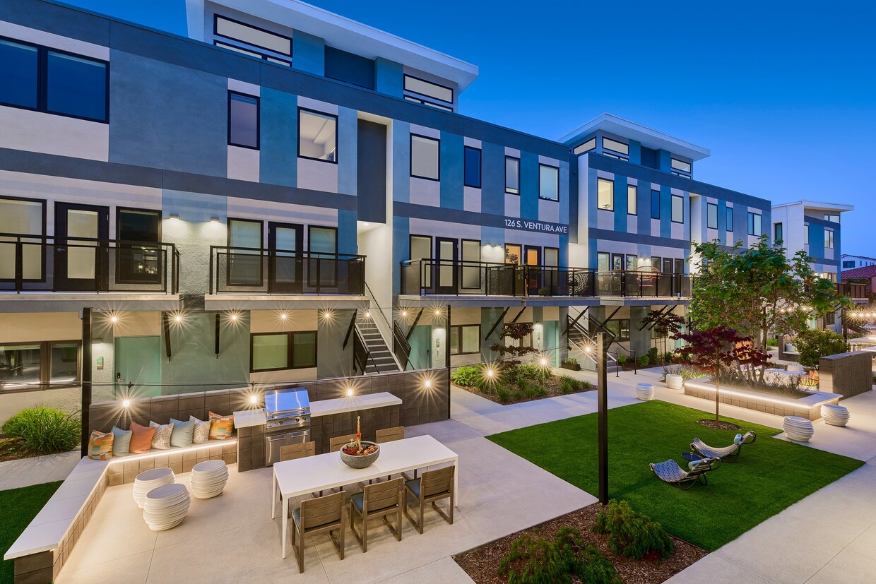 Modern multi-story building exterior at twilight, featuring blue and gray geometric color blocking, second-floor balconies with black railings, illuminated pathways, a landscaped courtyard with turf, string lights, and an outdoor seating and dining area with a built-in grill.