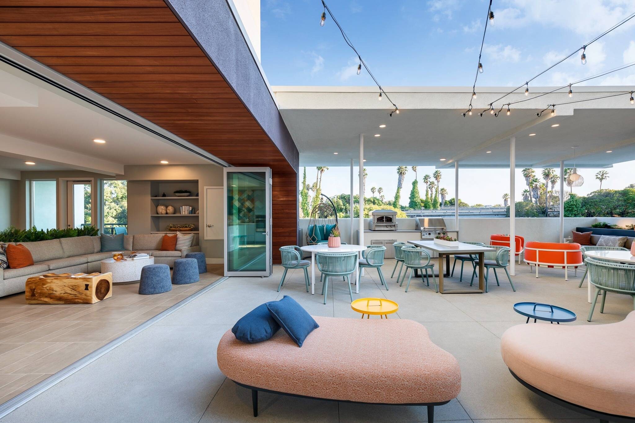 Modern outdoor rooftop patio featuring a peach-colored kidney-shaped daybed with blue throw pillows, string lights overhead, a round dining table with blue-green woven chairs, an outdoor kitchen and dining setup in the background, a hanging chair, and sliding glass doors opening into an indoor living space with wood ceiling accents.