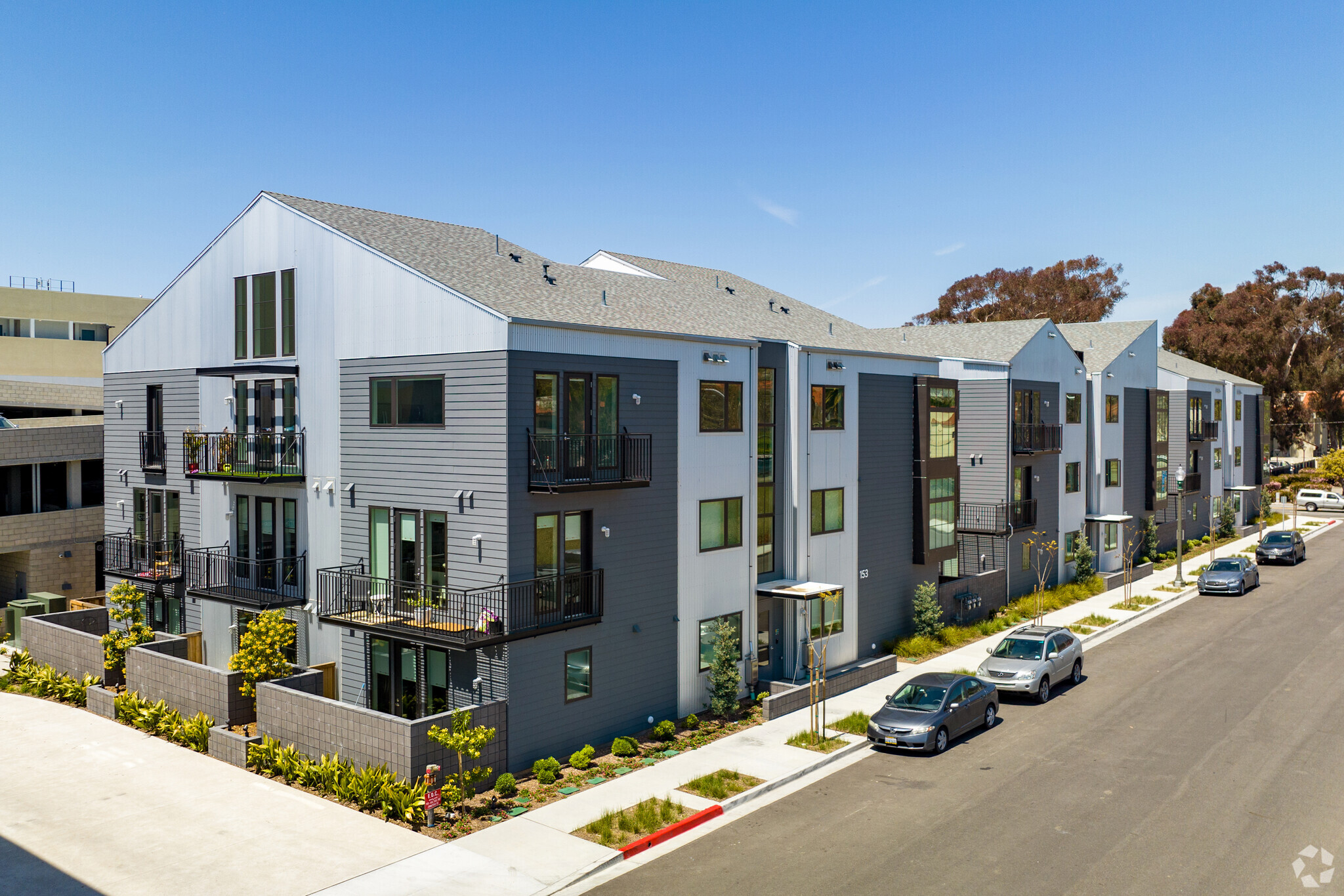 Modern multi-story apartment building exterior featuring a mix of gray siding, white vertical metal panels, black window trims, multiple balconies with black railings, a shingle roof, and street-level landscaping along a sidewalk with parked cars.