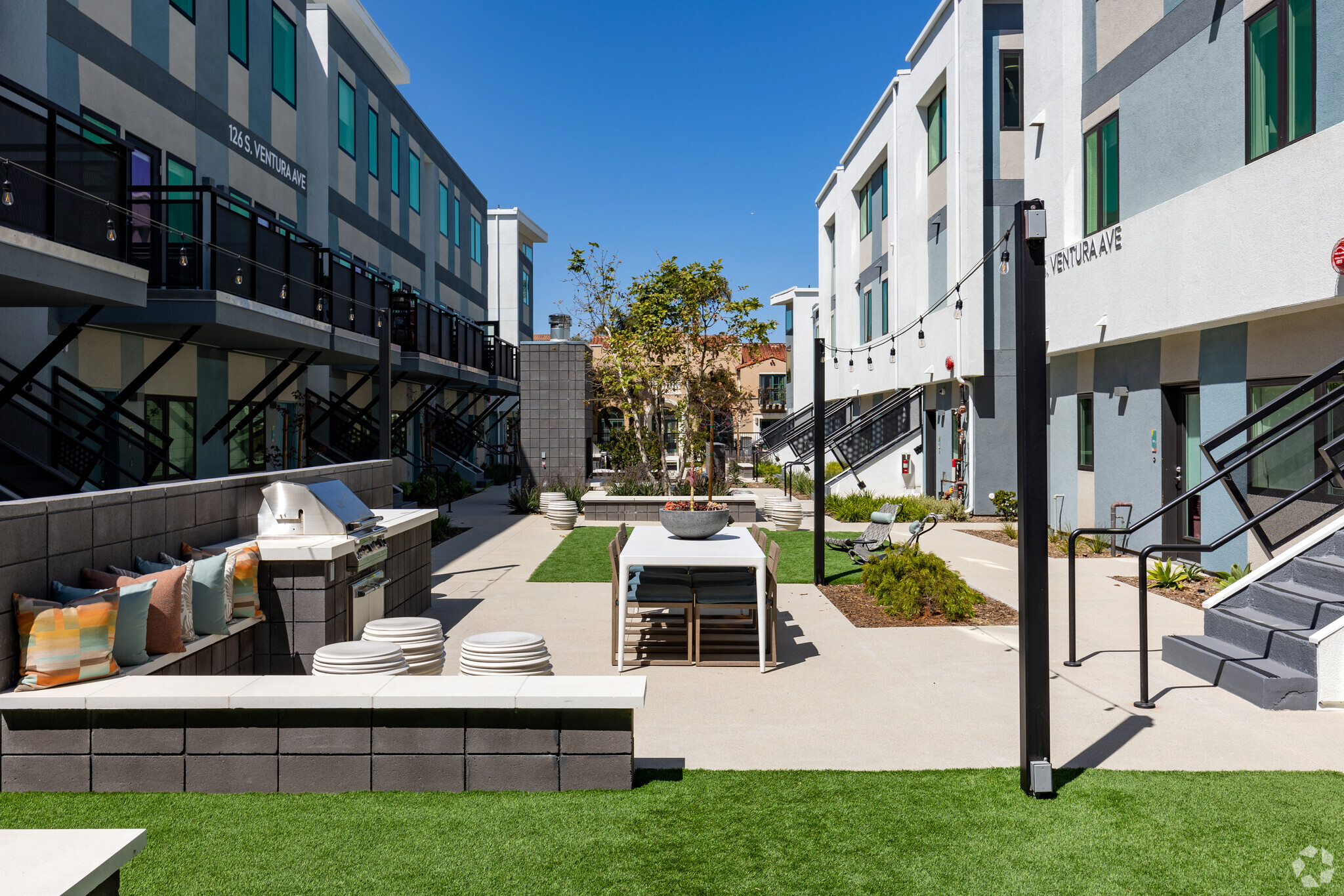 Modern courtyard space nestled between multi-story buildings, featuring a central dining table and chairs, artificial turf patches, string lights, and an outdoor kitchen area in the foreground.