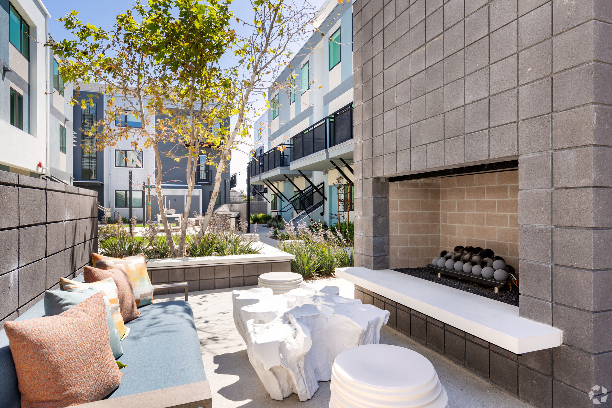 Modern outdoor courtyard space featuring a block-tile fireplace, a white carved wood-root coffee table, matching round side tables, a sofa with a blue cushion, and a landscaped area with trees and plants in front of multi-story buildings with balconies.