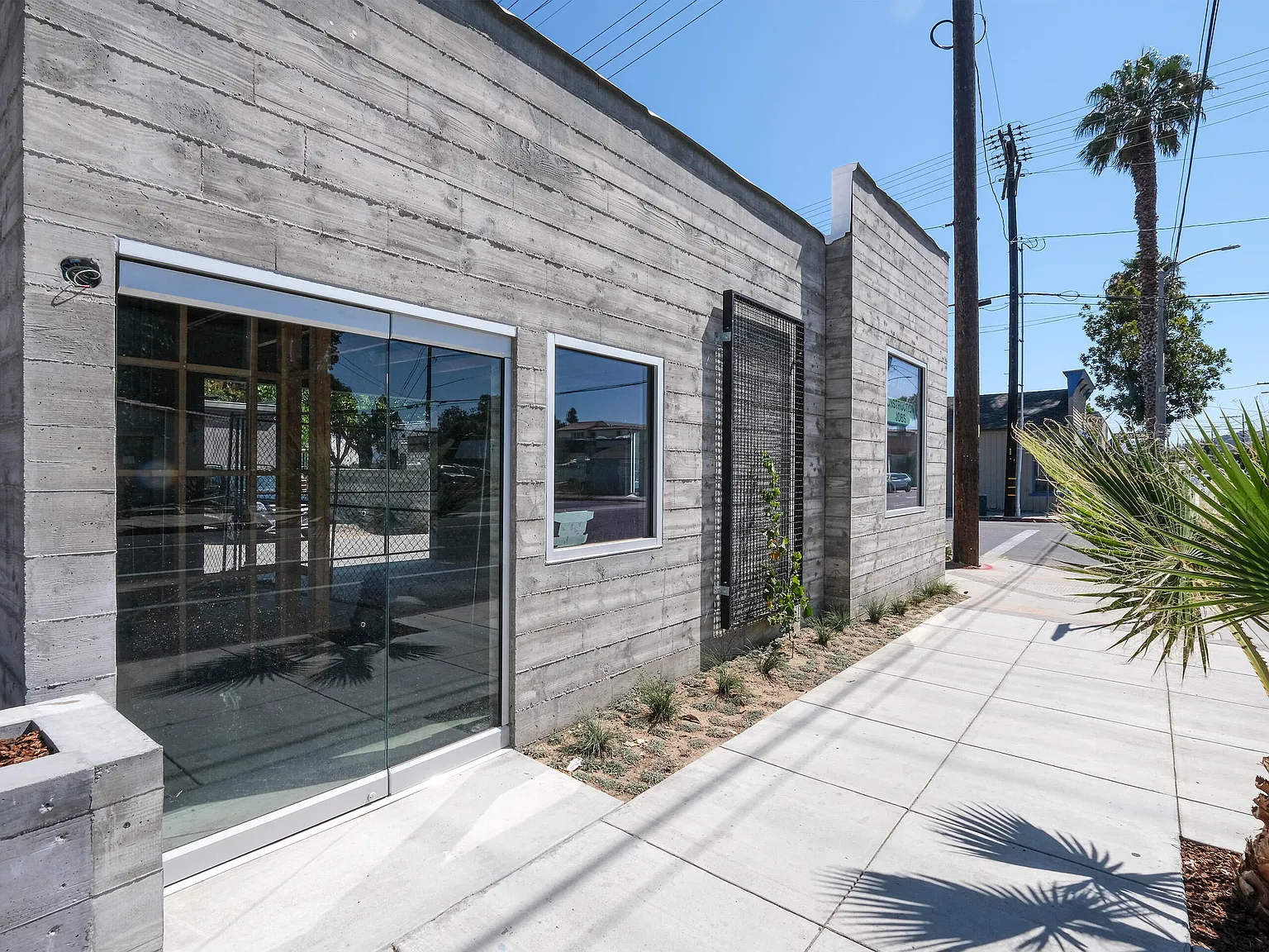 Modern concrete building exterior featuring board-formed concrete walls, large glass sliding doors, a square window, a black metal trellis with climbing greenery, and a sidewalk with landscaping and palm trees in the foreground.