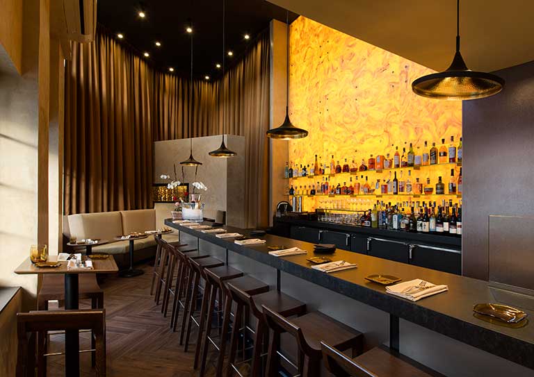 Luxurious bar and restaurant interior featuring a long dark countertop bar with matching stools, a striking backlit yellow onyx-style feature wall displaying rows of liquor bottles on glass shelves, and modern pendant lighting. The space is further enhanced by tall draped bronze curtains, booth seating along the left wall, and small dining tables set up for guests.