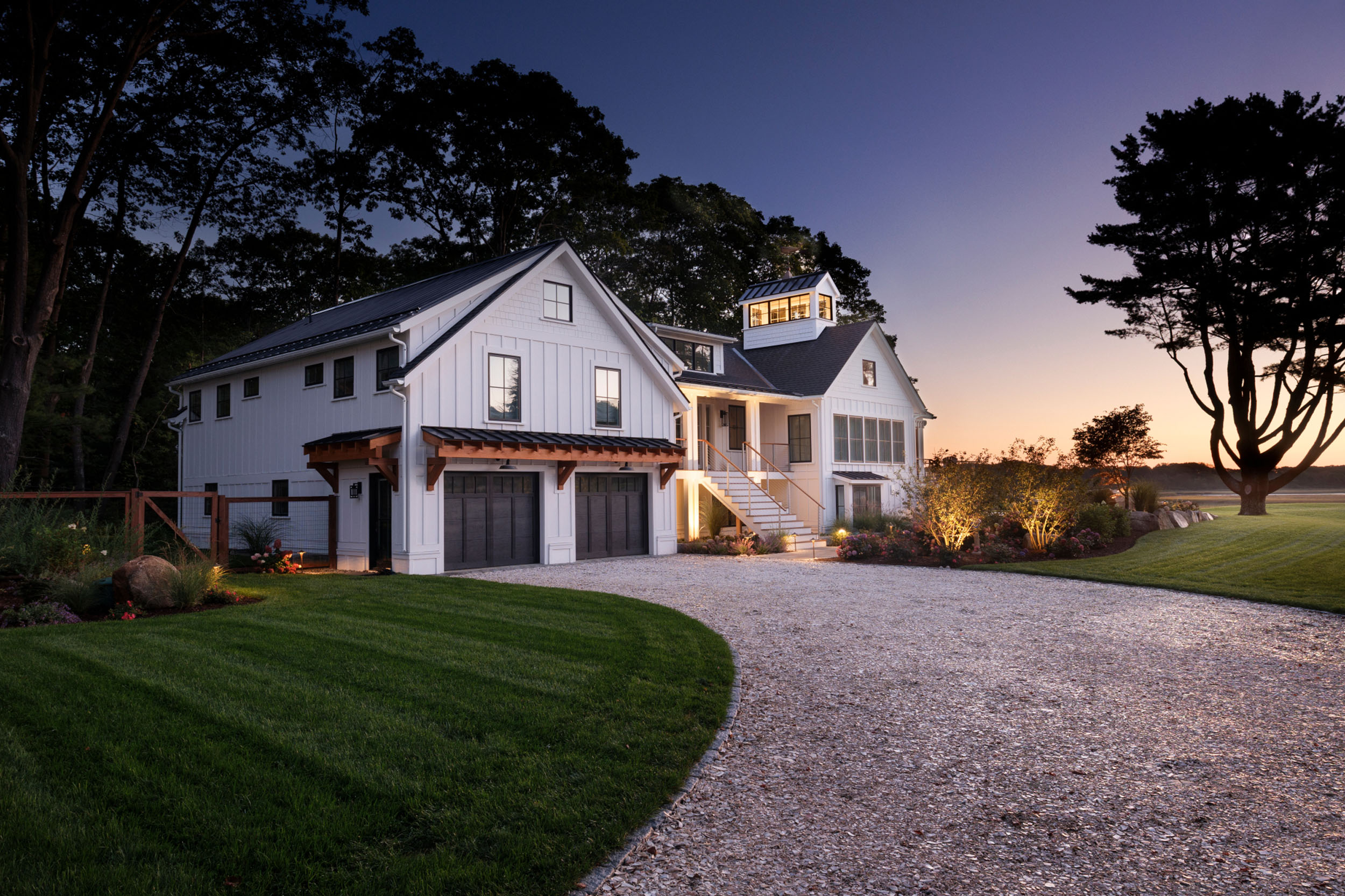 Custom modern farmhouse architecture featuring white board-and-batten siding, a timber-accented garage, illuminated entryway stairs, and a gravel driveway at twilight