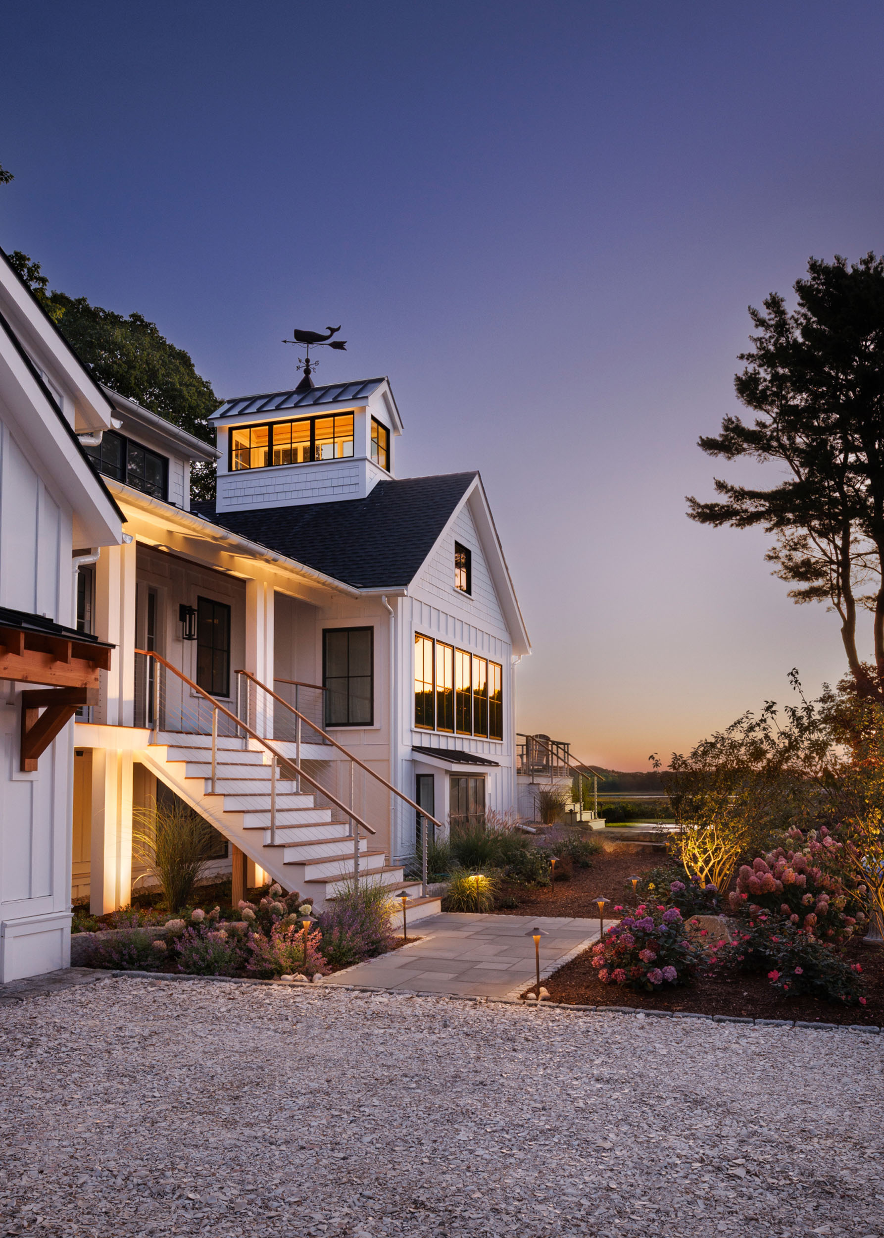 White coastal modern home at dusk featuring vertical siding, an exterior staircase leading to an upper deck, a lit cupola with a weathervane, a gravel driveway, and warm landscape lighting highlighting surrounding flower beds and trees
