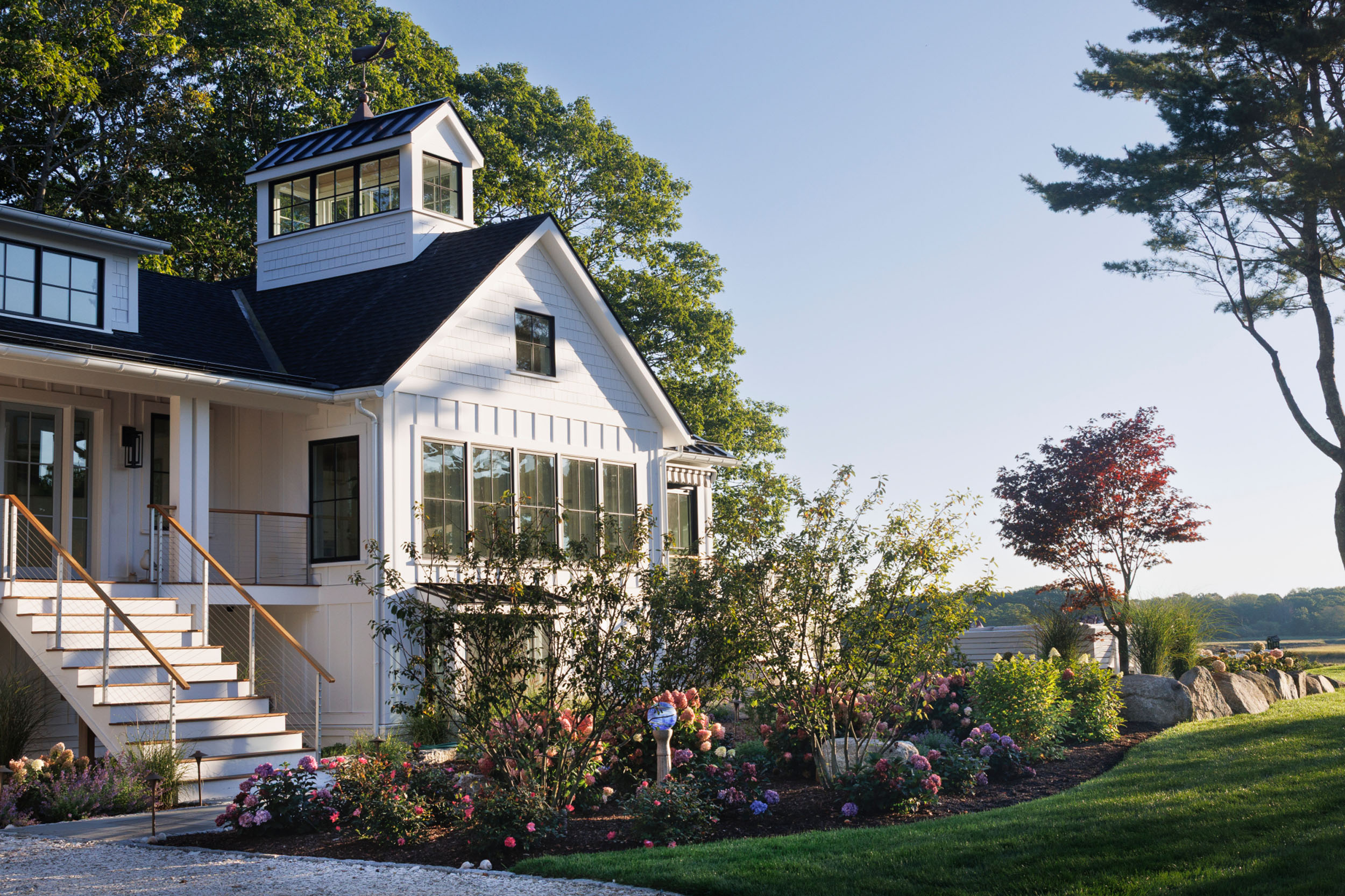 White coastal modern home exterior featuring vertical board-and-batten siding, shake gables, a black shingle roof with a cupola and weathervane, large multi-pane windows, and lush surrounding garden beds with flowering shrubs.