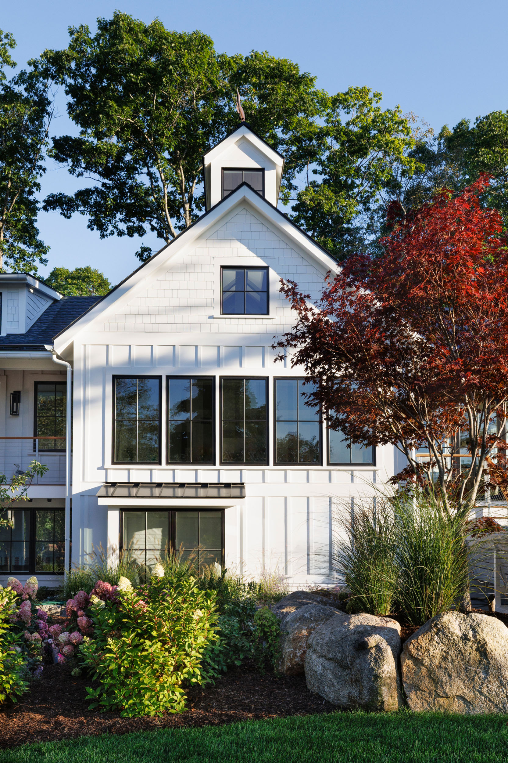 White coastal modern home exterior featuring vertical board-and-batten siding, shake gables, a black shingle roof with a cupola and weathervane, large multi-pane windows, and lush surrounding garden beds with flowering shrubs.