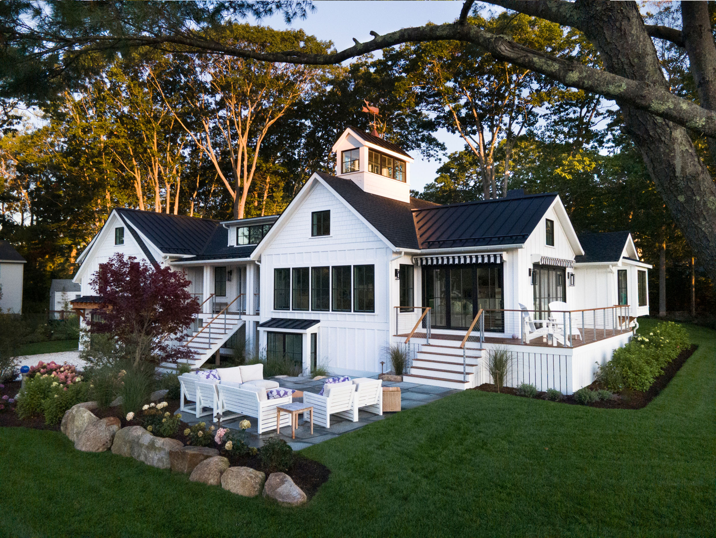 White coastal modern home exterior featuring vertical board-and-batten siding, shake gables, a black shingle roof with a cupola and weathervane, large multi-pane windows, and lush surrounding garden beds with flowering shrubs.