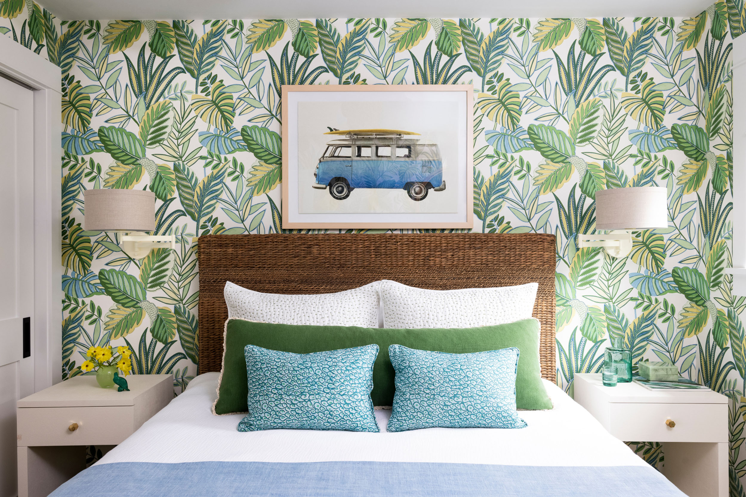 Eclectic bedroom detail featuring a woven rattan headboard, bold tropical leaf wallpaper, a framed art print of a blue vintage surf van, and layered bedding with green and blue patterned pillows.