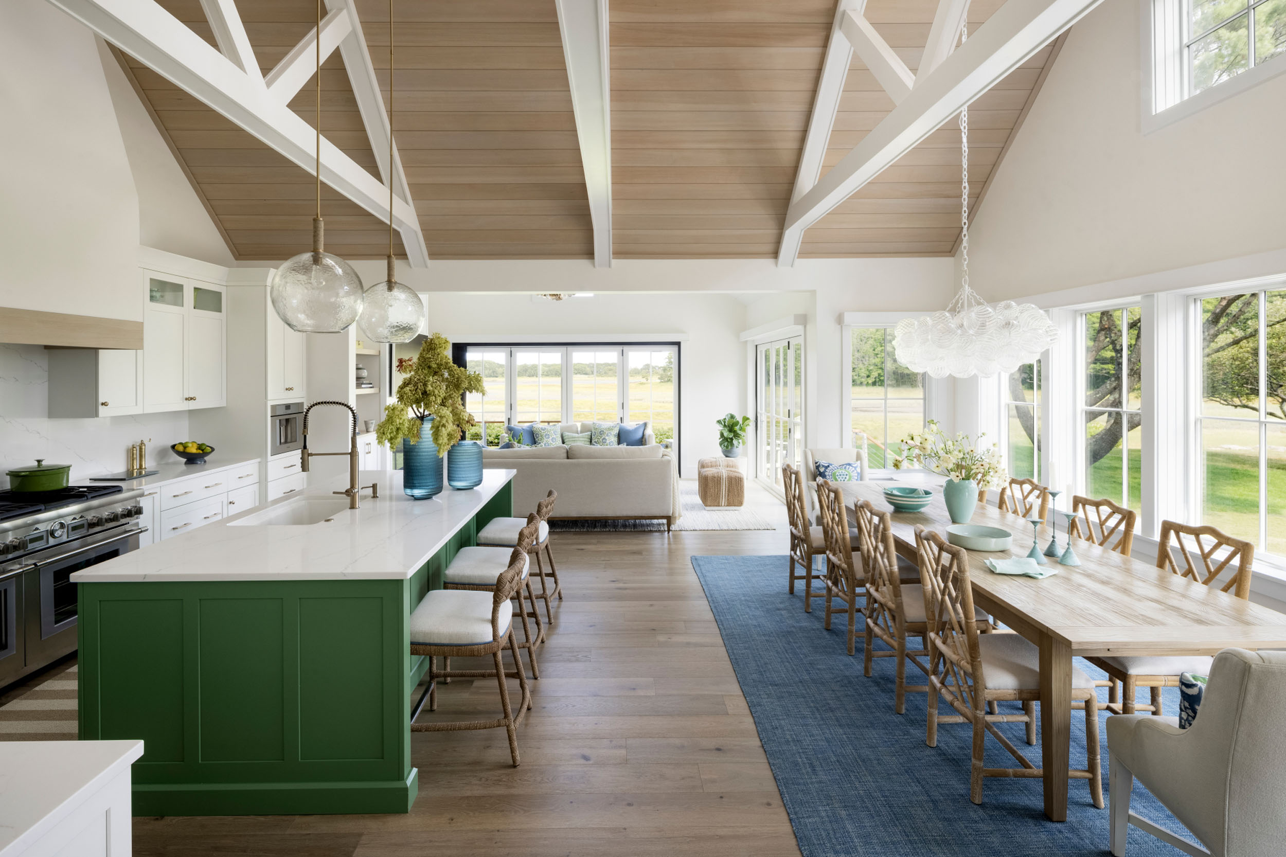Open-concept living, kitchen, and dining area featuring a vaulted wood-plank ceiling with white beams, a green kitchen island with white quartz countertops and woven barstools, a blue dining rug, and a light-filled living space opening to views outside.