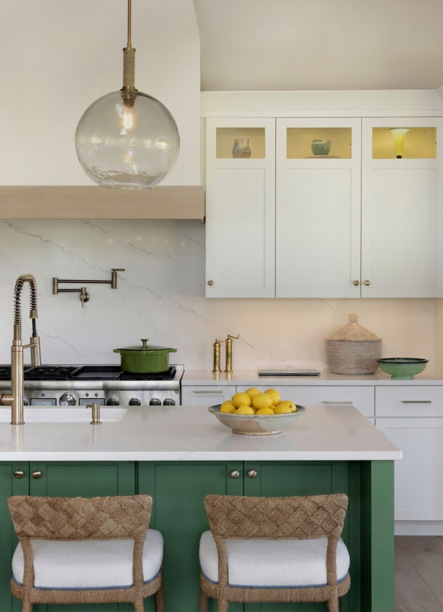 Bright modern kitchen featuring a green island base with white countertops and woven barstools, white upper and lower cabinetry, a marble backsplash, a pot filler above the range, and a large glass globe pendant light overhead.
