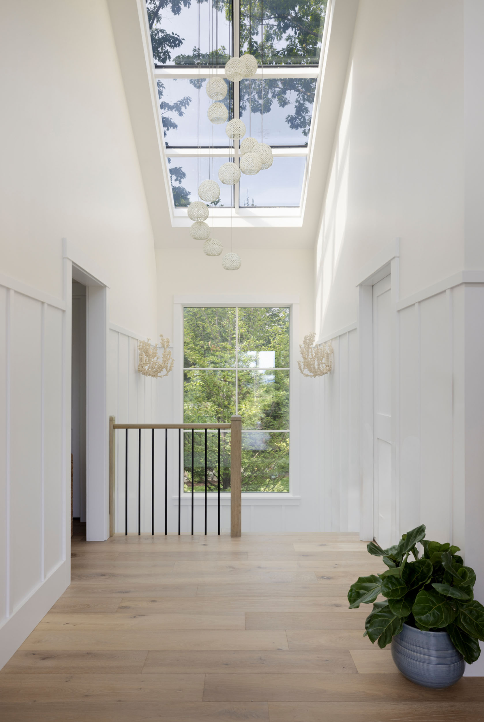 Bright modern upstairs hallway featuring white board-and-batten wainscoting, light hardwood floors, a large skylight overhead with a cascading cluster of textured globe pendant lights, a staircase railing with black spindles, and a potted plant in the corner.