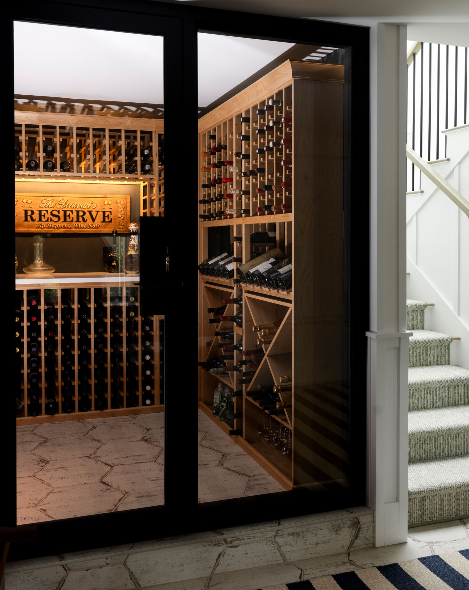 Custom enclosed wine cellar featuring extensive light wood racking, a lighted display sign, hexagonal floor tiling, and a modern glass door next to a staircase with patterned carpeting.
