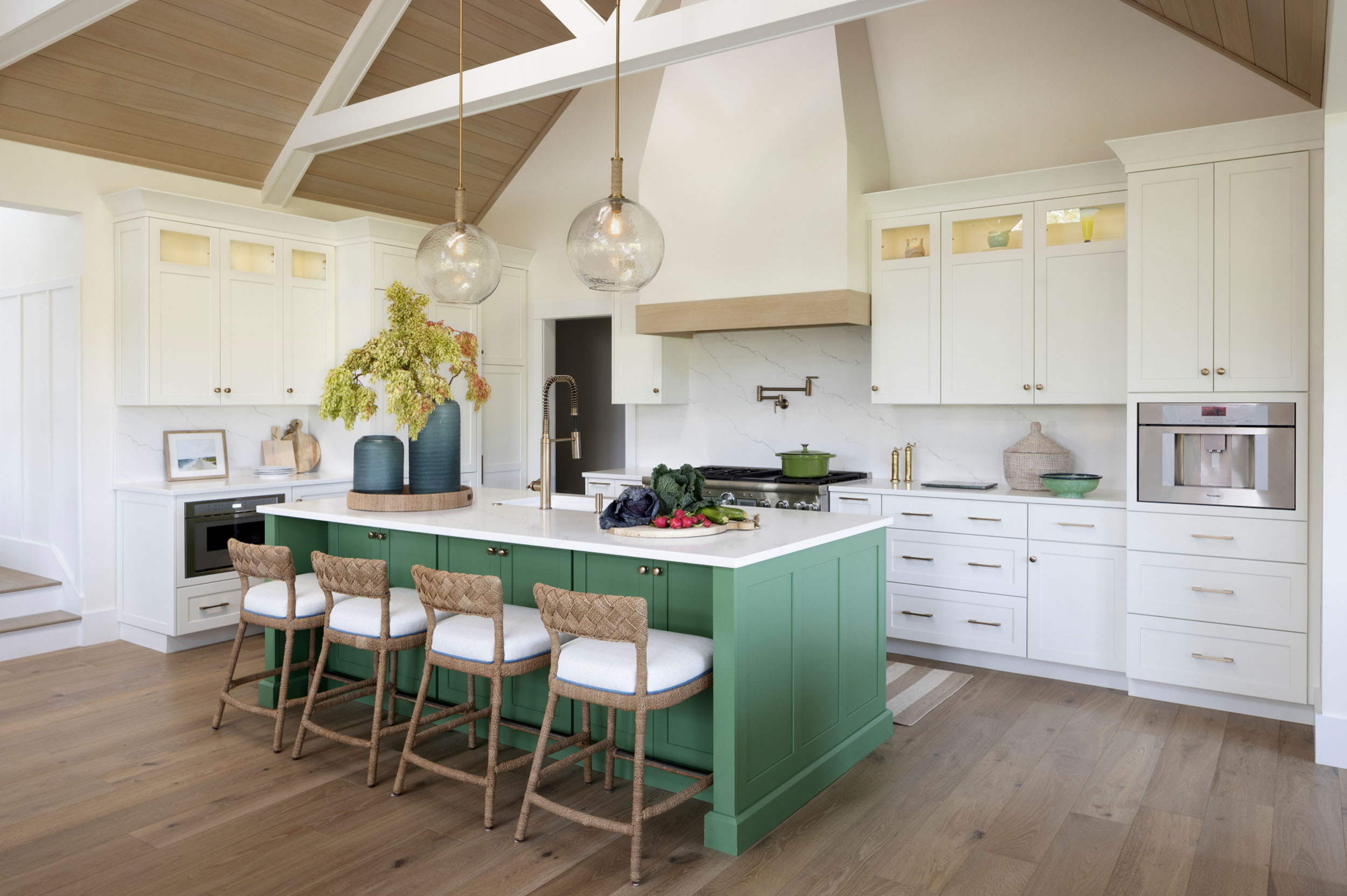 Bright, spacious modern kitchen featuring a bold green central island with white quartz countertops and four woven rattan barstools. The space is accented with white perimeter cabinetry, lit glass-front upper cabinets, a marble backsplash with a pot filler over the range, and large glass globe pendant lights hanging from a vaulted wooden ceiling.