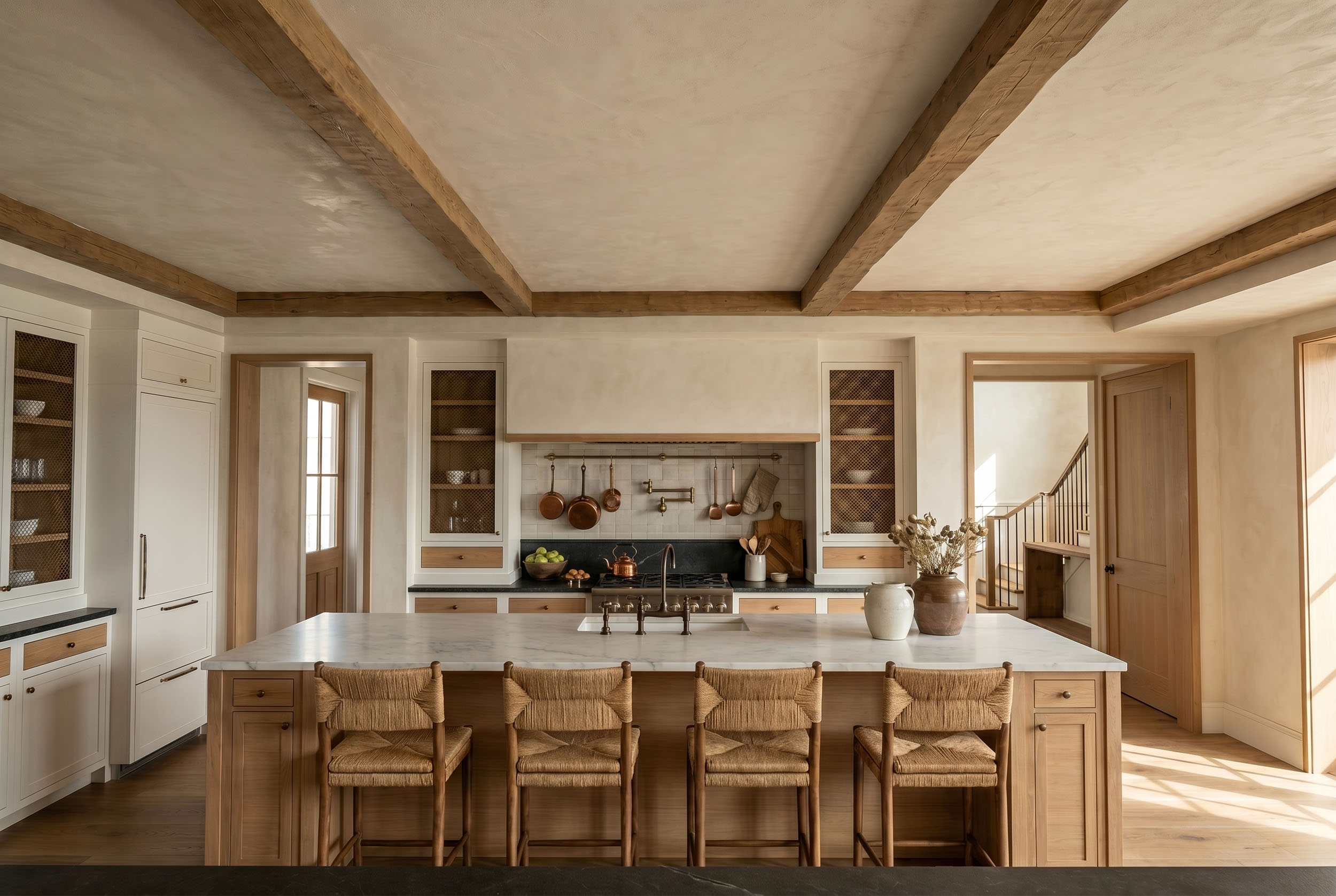 Warm modern kitchen interior featuring exposed wood ceiling beams, a marble island with woven barstools, mesh-front display cabinetry, and a plastered range hood over copper cookware accents
