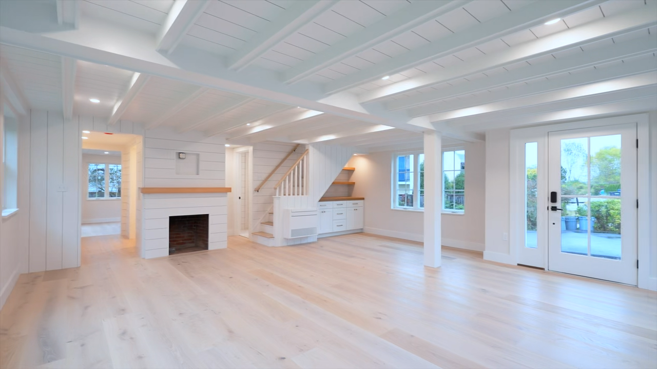 Bright and airy modern interior featuring light wood flooring, white shiplap walls and a beamed ceiling, a staircase with a white railing, built-in lower cabinetry with open shelving beneath the stairs, a fireplace, and large multi-pane windows.