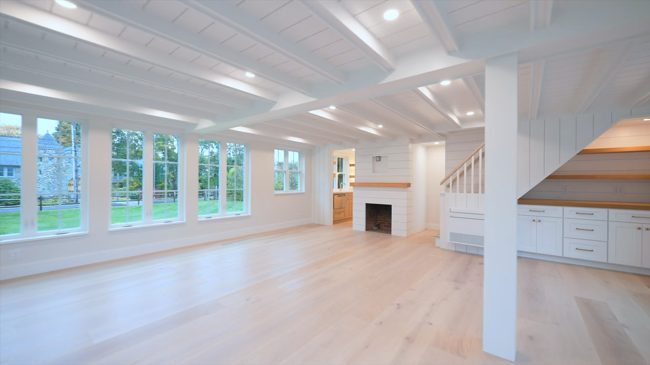 Bright and airy modern interior featuring light wood flooring, white shiplap walls and a beamed ceiling, a staircase with a white railing, built-in lower cabinetry with open shelving beneath the stairs, a fireplace, and large multi-pane windows.