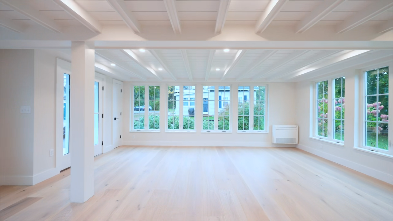 Bright and airy modern interior featuring light wood flooring, white shiplap walls and a beamed ceiling, a staircase with a white railing, built-in lower cabinetry with open shelving beneath the stairs, a fireplace, and large multi-pane windows.