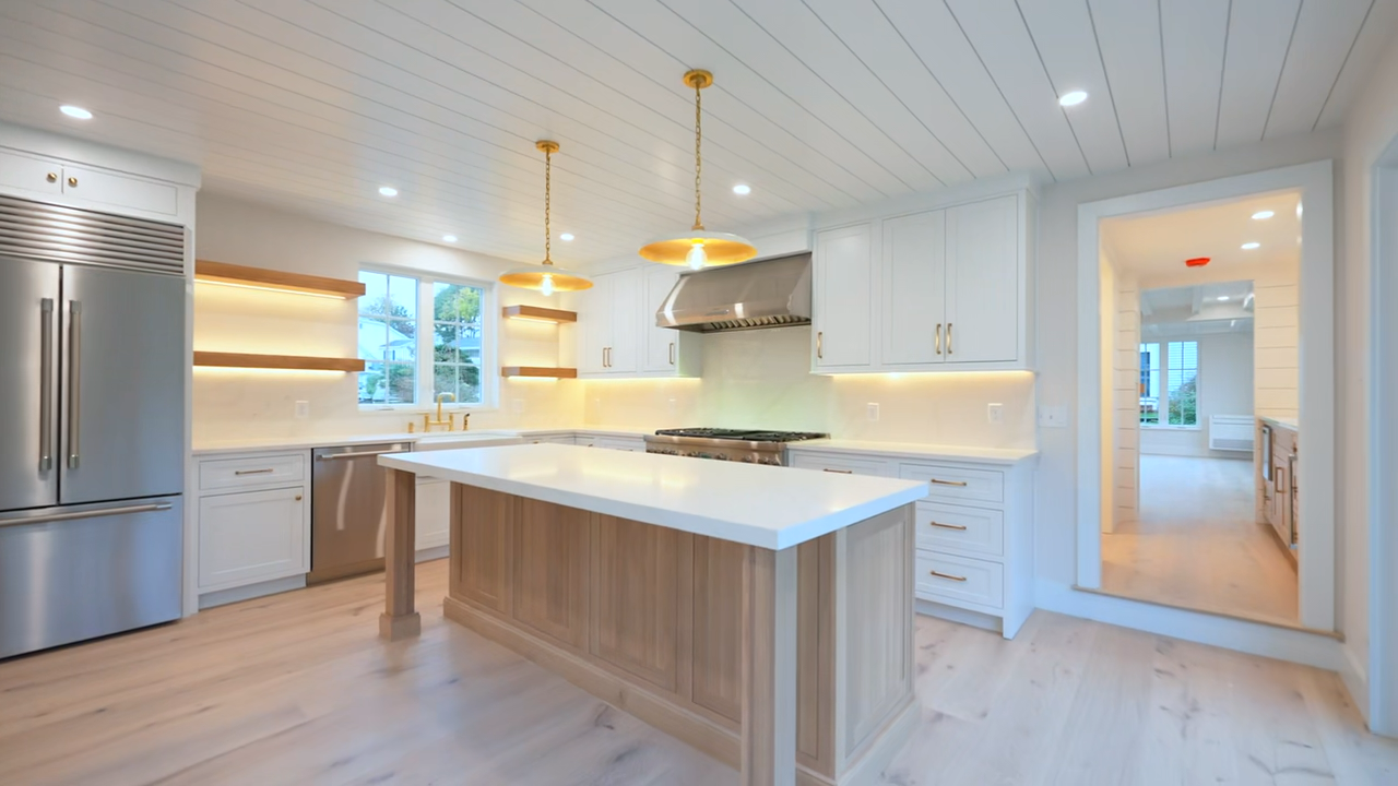 Modern kitchen area featuring a white countertop, an undermount sink, a sleek gold faucet, light wood lower cabinets, white horizontal shiplap walls, and illuminated floating wooden shelves next to a multi-pane window.