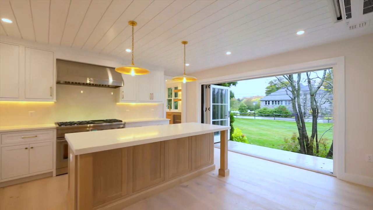 Modern kitchen featuring a white center island with a light wood base, white perimeter cabinetry, a matching white countertop, illuminated floating wooden shelves, a stainless steel range hood, a shiplap ceiling, and two gold pendant lights.