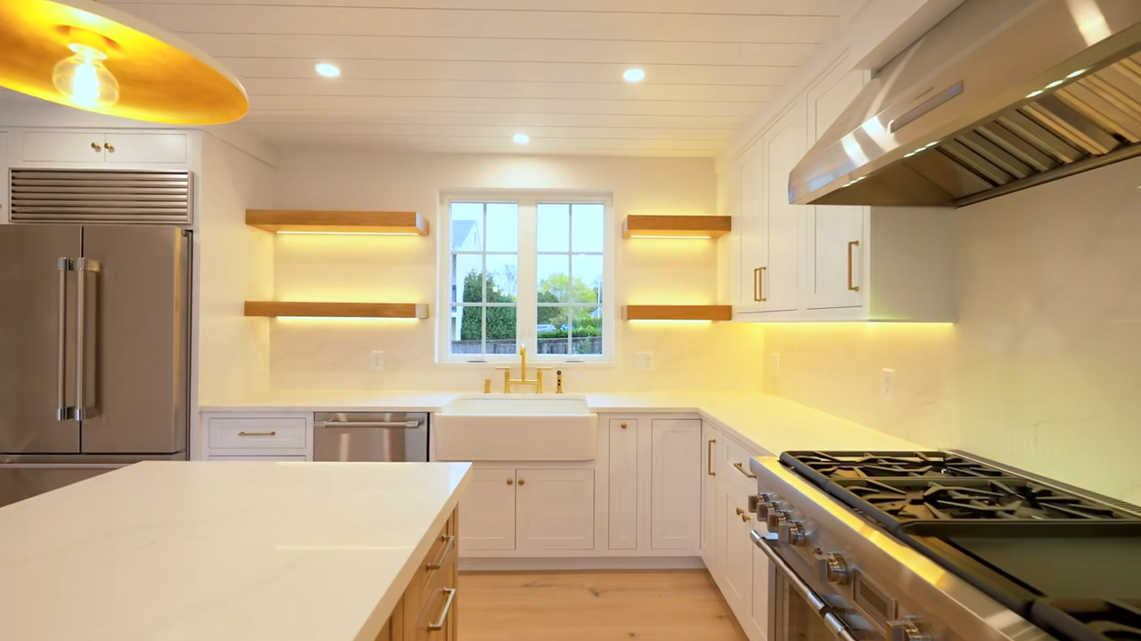 Modern kitchen featuring a white center island with a light wood base, white perimeter cabinetry, a stainless steel range hood, a shiplap ceiling, gold pendant lights, and a large open sliding glass door leading to an outdoor view of a lawn and trees.