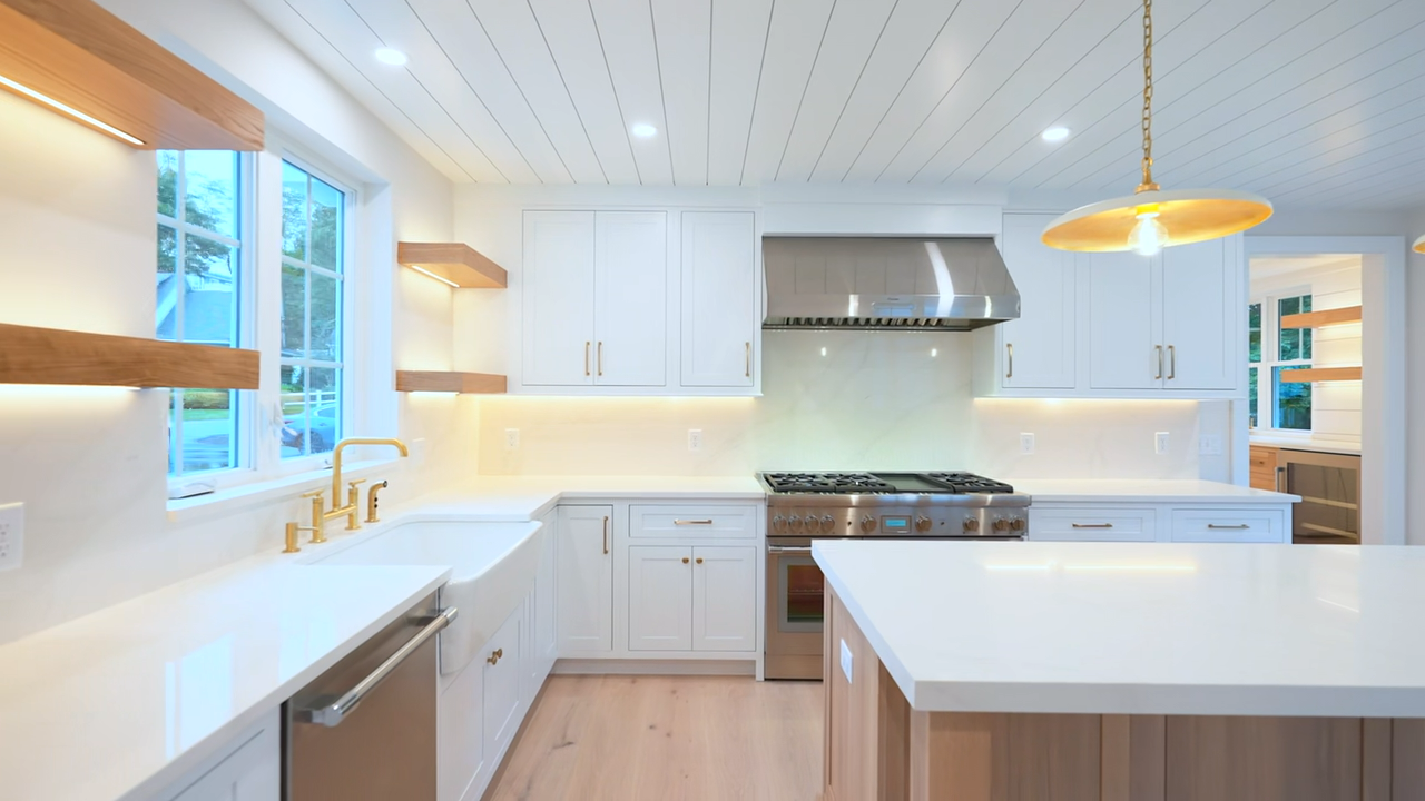 Modern kitchen featuring white perimeter cabinetry, a large white farmhouse sink beneath a multi-pane window, illuminated floating wooden shelves, a stainless steel range and hood, white countertops, and a stainless steel refrigerator.