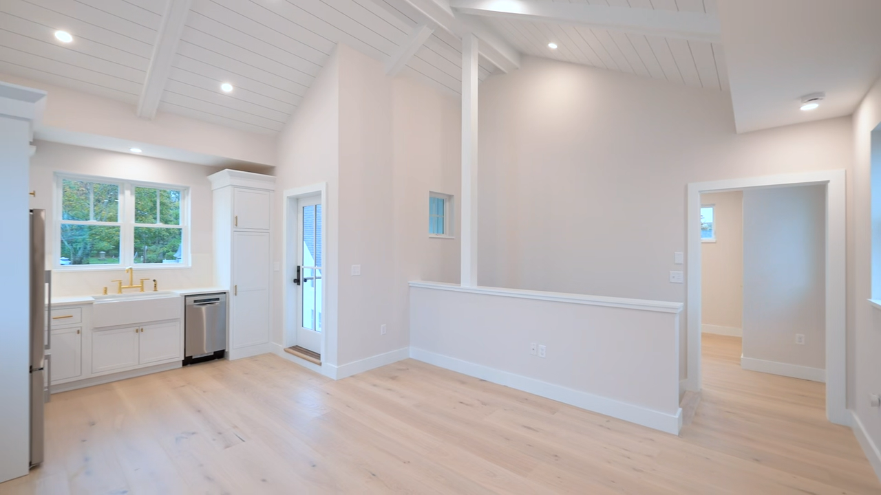 Bright and airy interior space featuring light wood flooring, white walls, a vaulted shiplap ceiling with exposed beams, a partial-height dividing wall, a doorway leading to an adjacent room, and a kitchen area with white cabinetry, a sink beneath a window, and a stainless steel dishwasher.