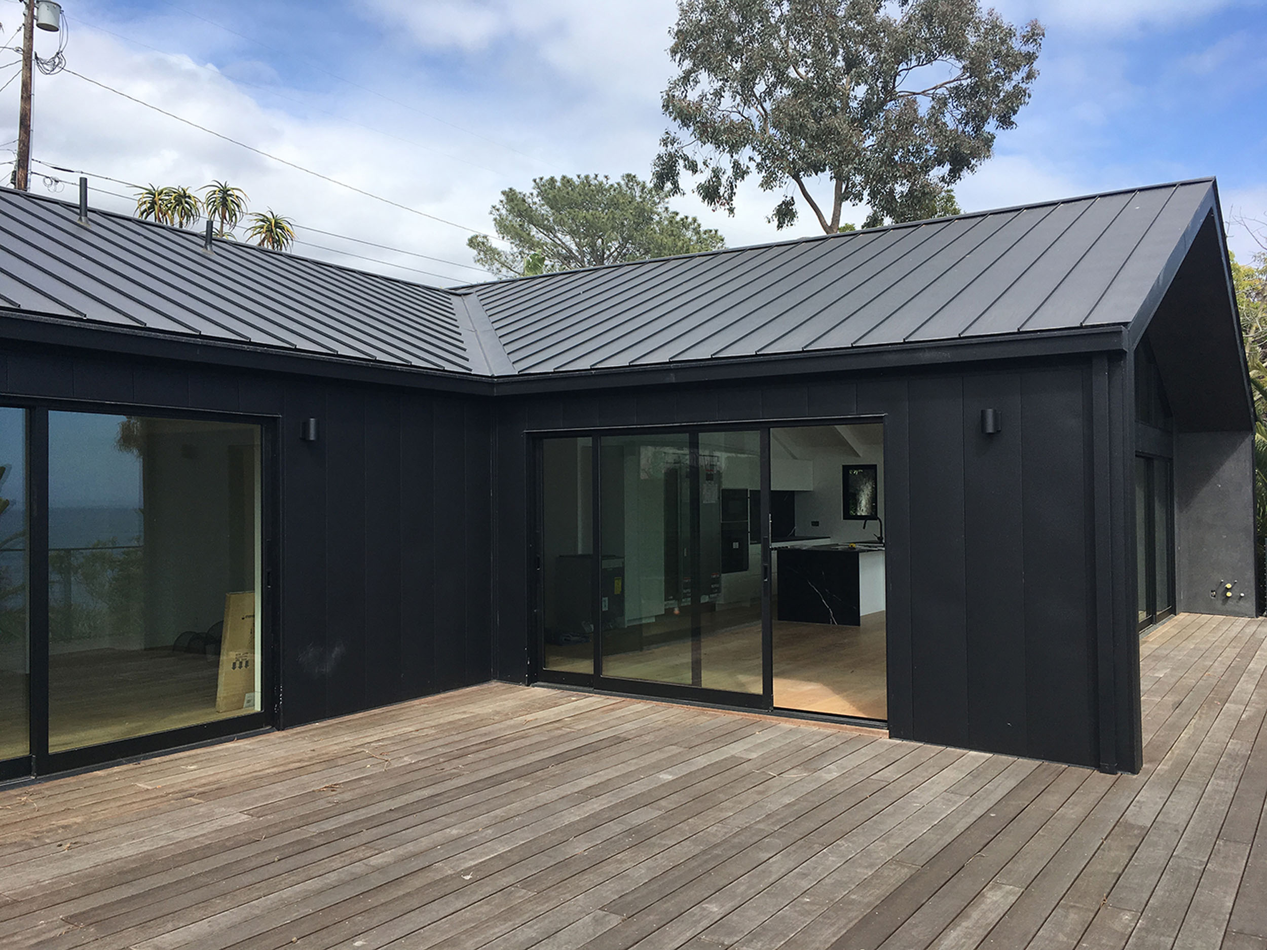 Modern single-story home exterior featuring dark vertical siding, a dark metal standing seam roof, large sliding glass doors, and a spacious wood deck.