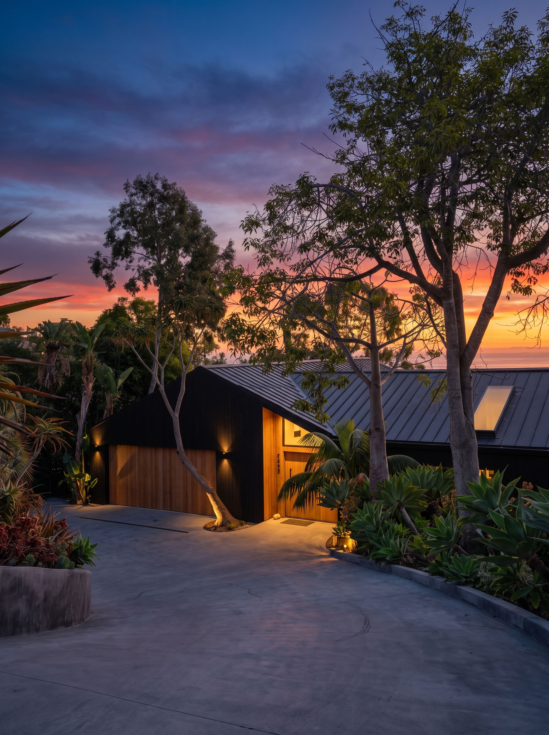Modern single-story home featuring black vertical siding, a dark standing-seam metal roof, warm wood garage and entry accents, and lush tropical landscaping at sunset overlooking the ocean