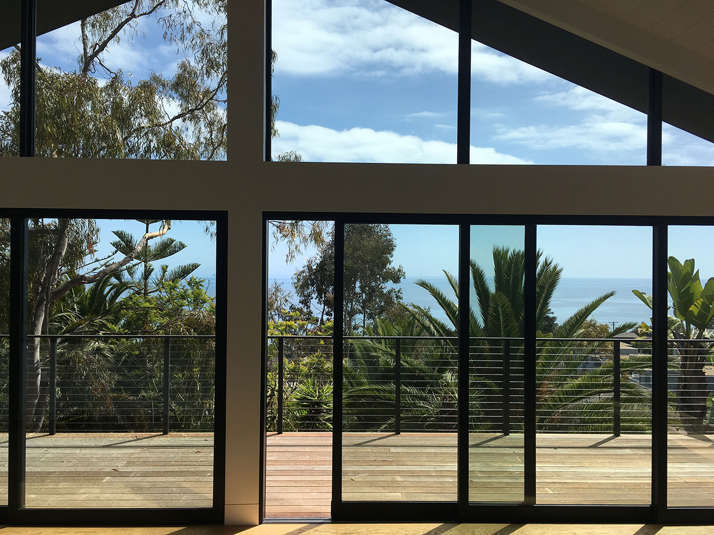 Large floor-to-ceiling windows and sliding glass doors with black framing, looking out over a balcony with a cable railing, lush tropical foliage, palm trees, and an open blue ocean view under a partly cloudy sky.