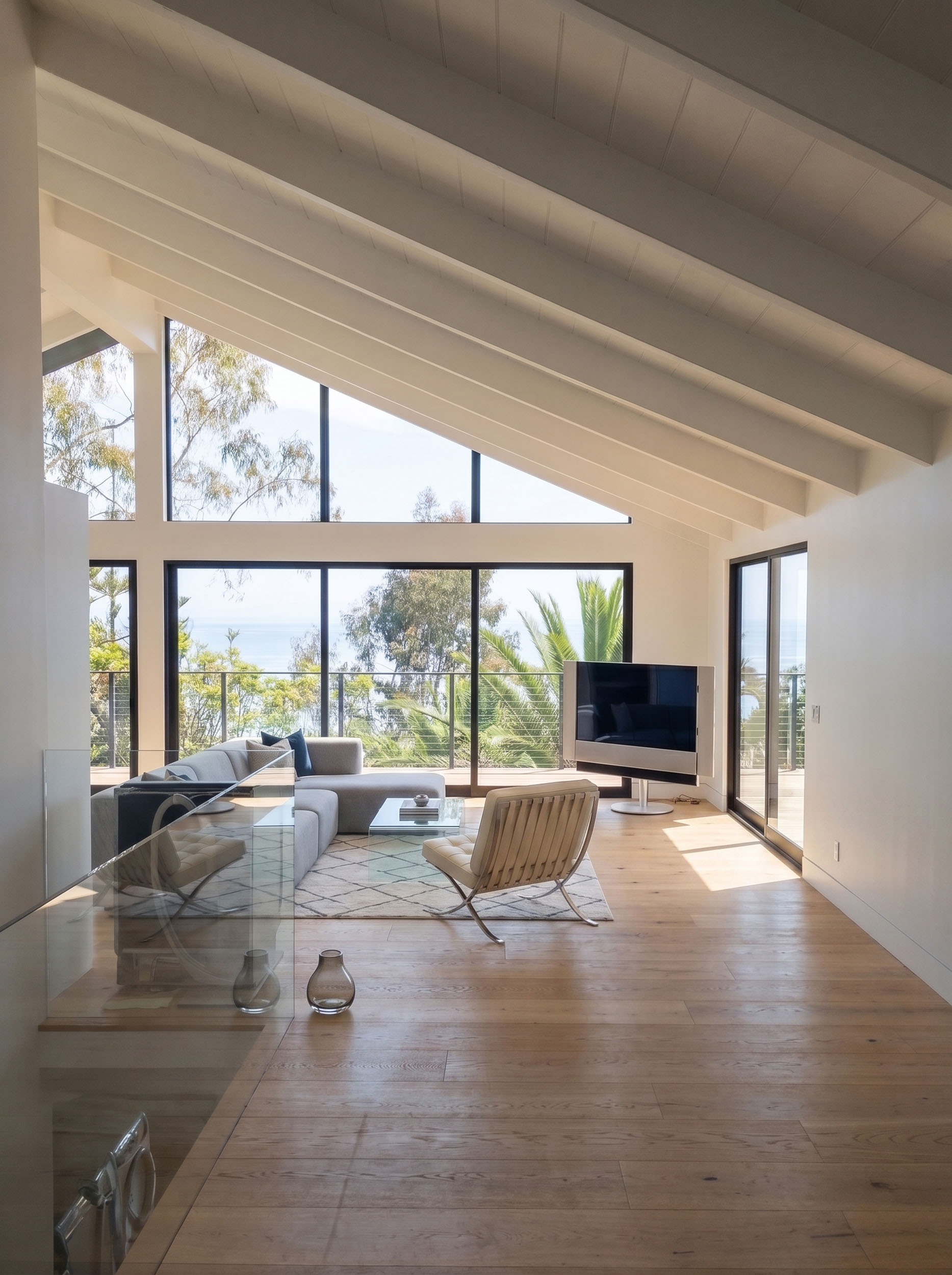 Bright and modern living room featuring light wood flooring, a vaulted ceiling with exposed white beams, and an expansive wall of windows and sliding glass doors that frame an ocean view and lush greenery. The seating area includes a neutral sectional sofa, a glass coffee table over a geometric rug, and a modern lounge chair, with a sleek television positioned on a stand near the window.
