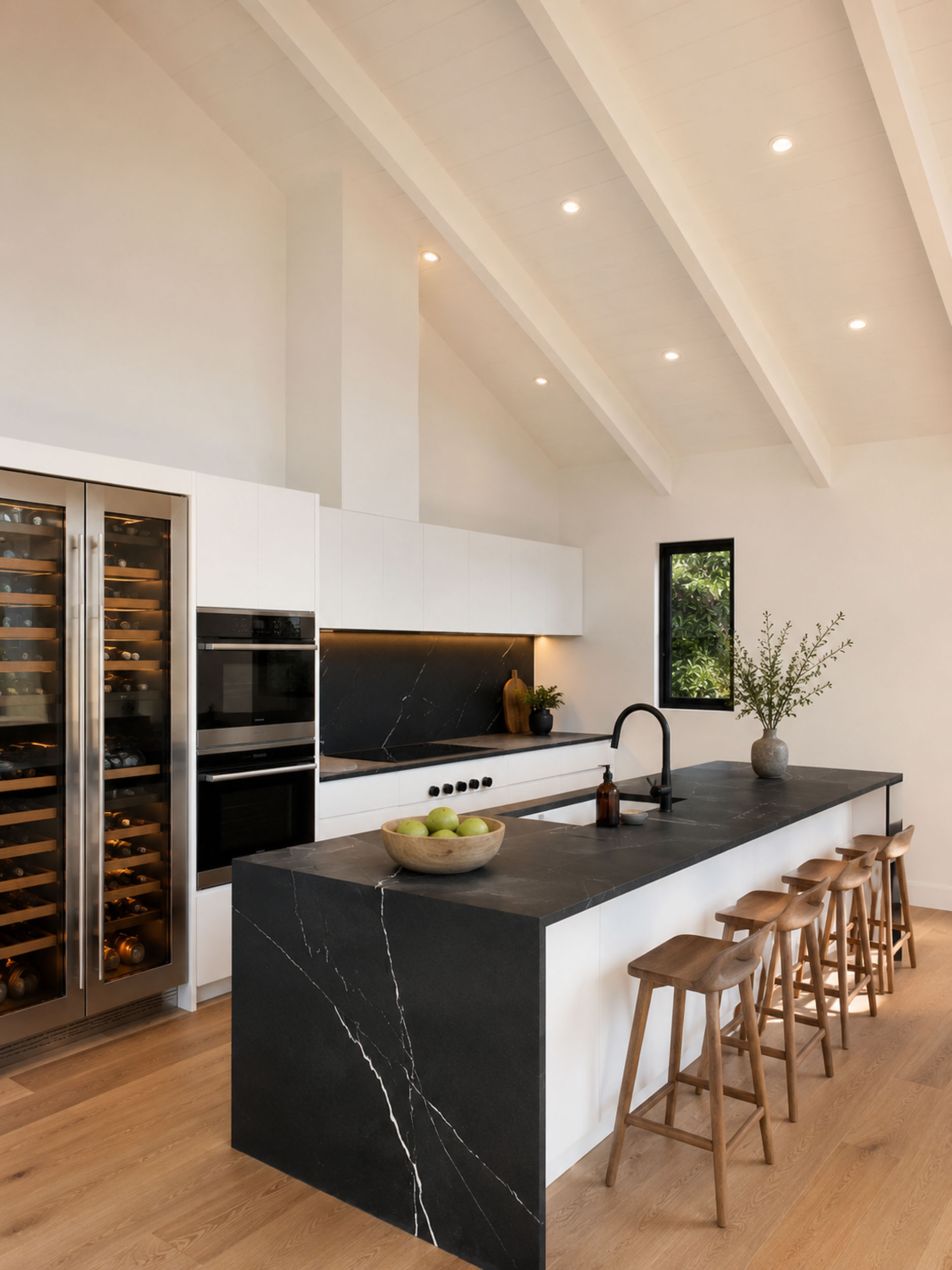 Modern kitchen featuring white perimeter cabinetry, dark marble-look countertops and a matching waterfall island, built-in double ovens, a stainless steel dual-zone wine refrigerator, a vaulted ceiling with exposed white beams, and a row of wooden barstools.