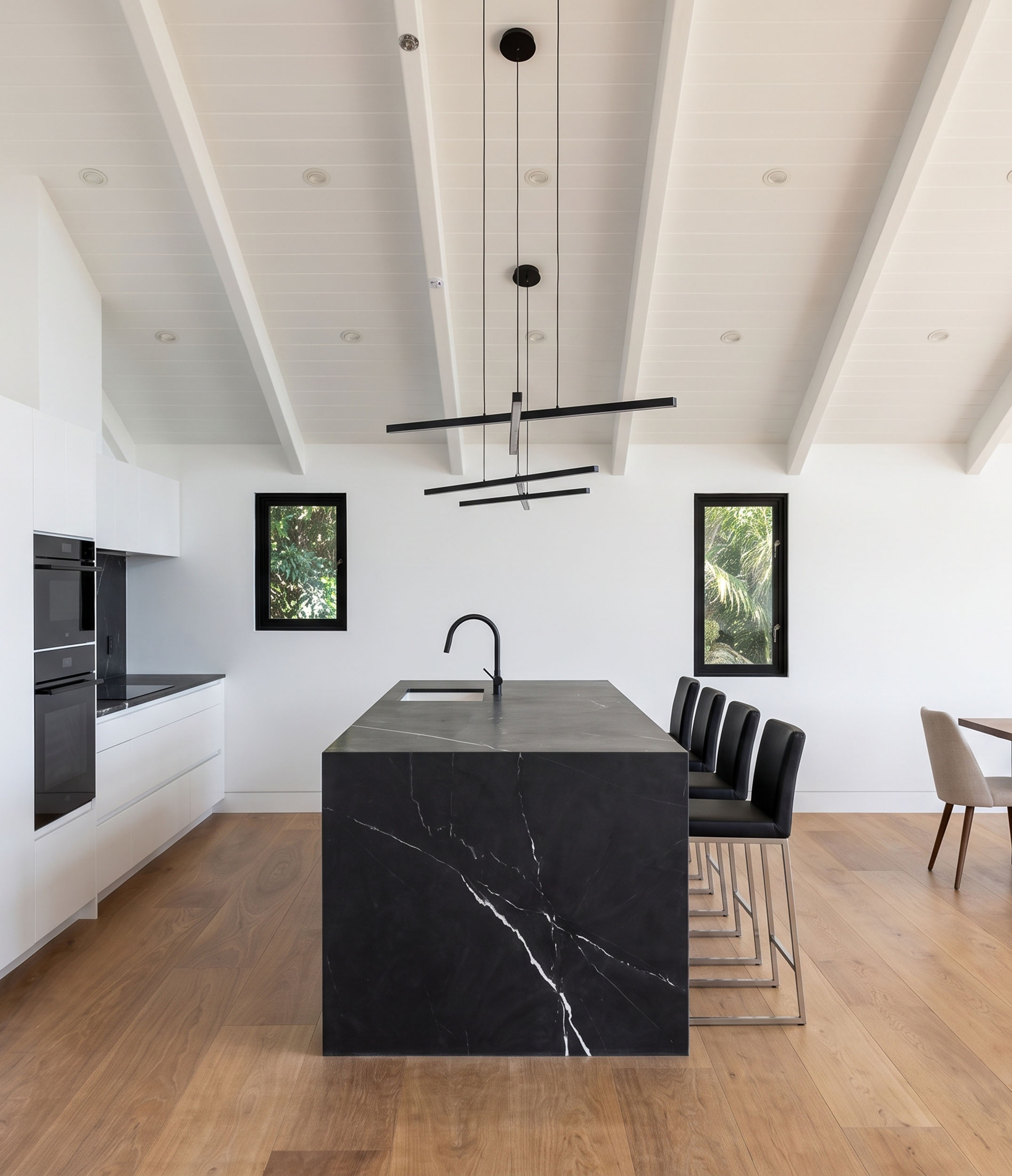 Modern kitchen interior featuring a striking dark marble waterfall island with white veining, sleek white perimeter cabinetry, built-in double ovens, light wood flooring, a vaulted ceiling with exposed white beams, two small black-framed windows, a modern linear black pendant light fixture, and black leather barstools.